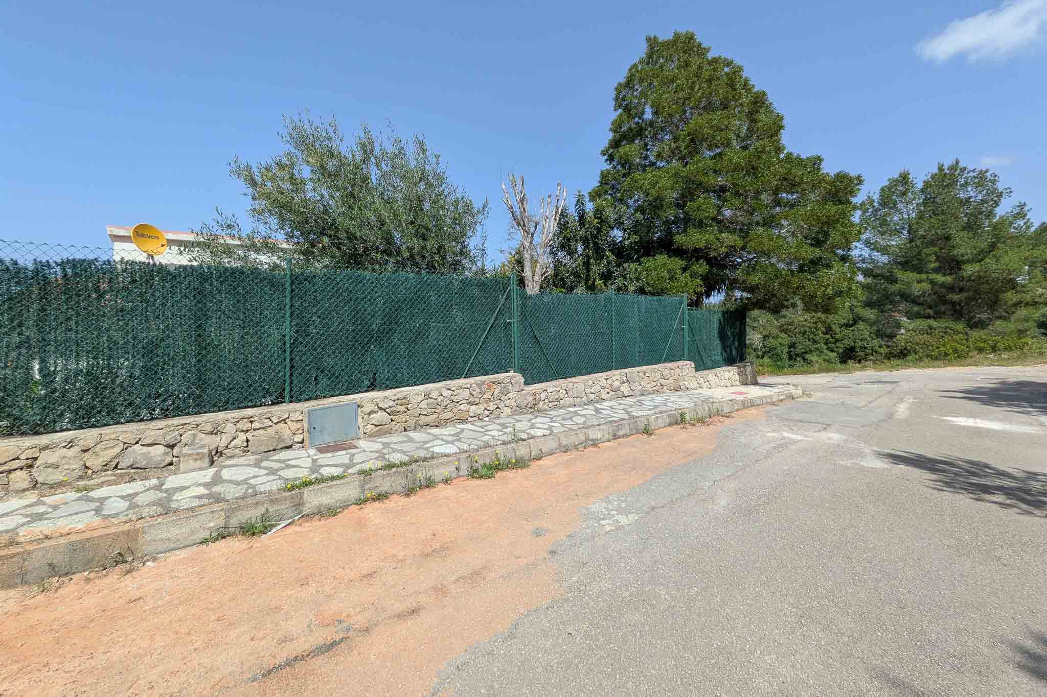 Fachada exterior con muro de piedra natural, vallado perimetral de ocultación y acera empedrada en una tranquila calle residencial de La Pedrera.
