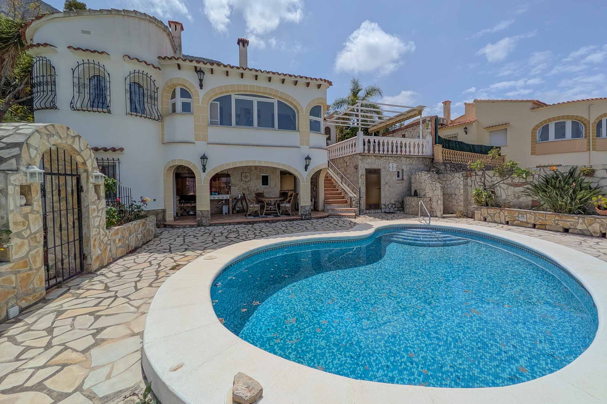 Piscina de gresite azul integrada en una terraza de piedra natural, con porche cubierto que alberga la cocina de verano y comedor exterior.