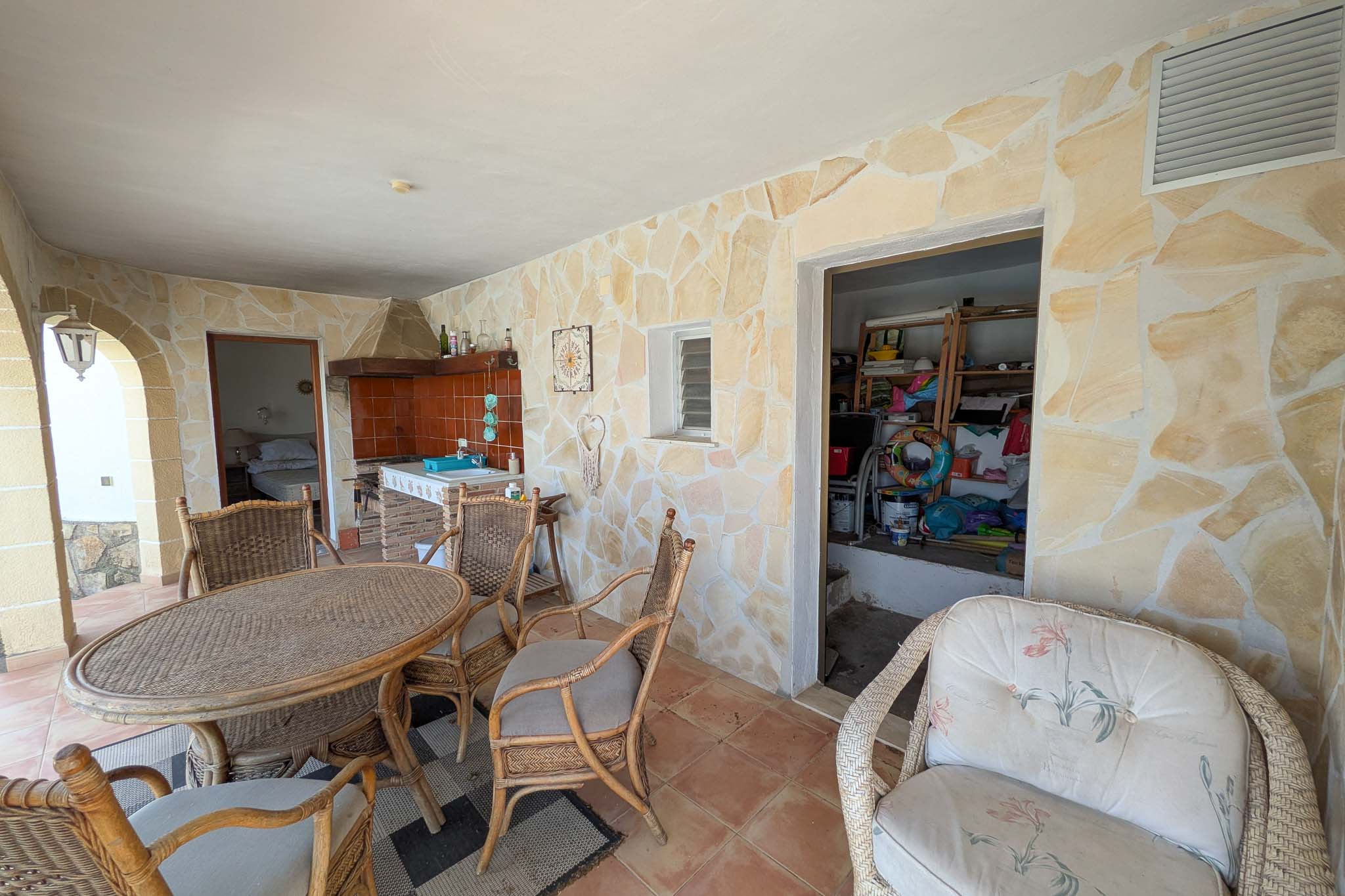 Terraza cubierta en planta baja con paredes de piedra natural, cocina de verano integrada y zona de comedor con mobiliario de mimbre junto al acceso al trastero.