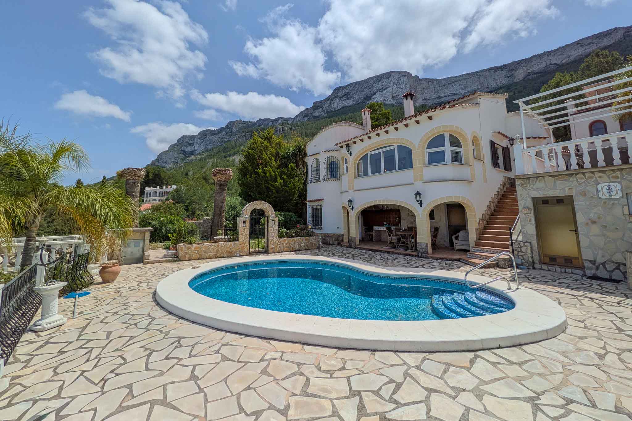 Vista exterior de la piscina de forma irregular rodeada de una terraza de piedra natural, con la fachada blanca de arcos y el monte Montgó al fondo.