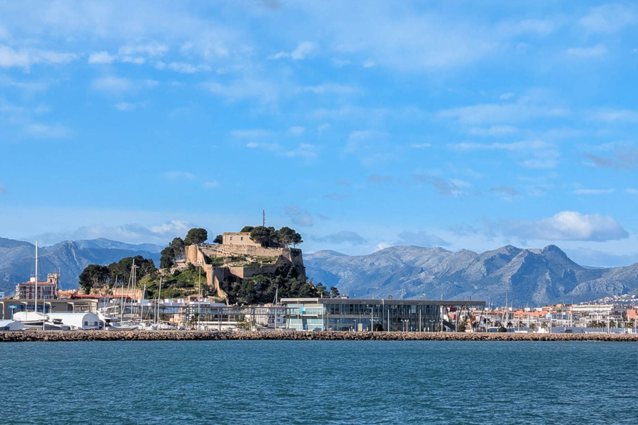 Vista panorámica del Castillo de Dénia y el puerto deportivo, destacando la arquitectura histórica sobre la colina y las montañas al fondo.