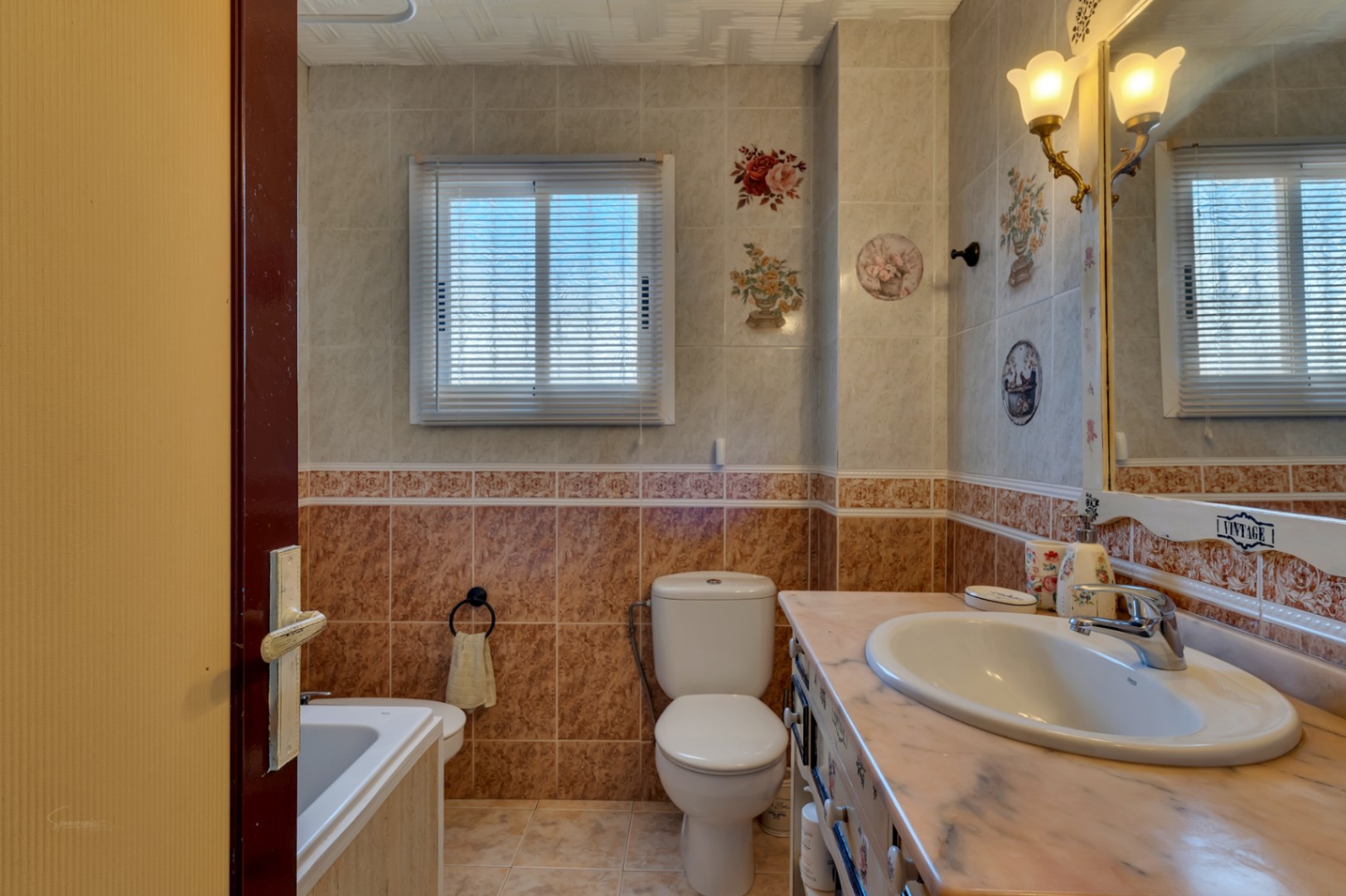 Baño completo con azulejos decorativos, encimera de mármol, espejo con apliques clásicos y ventana con persiana que aporta luz natural.