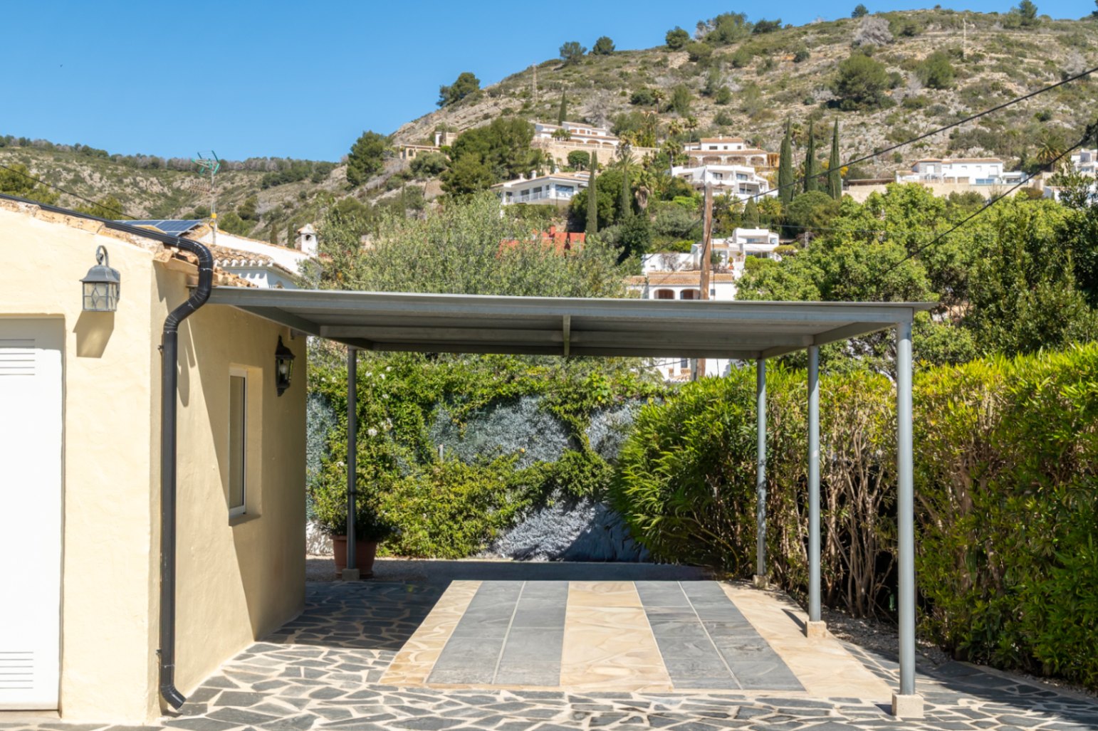 Carport metálico sobre suelo de piedra natural y pizarra, situado junto a la fachada principal con vistas a la ladera arbolada de Jávea.