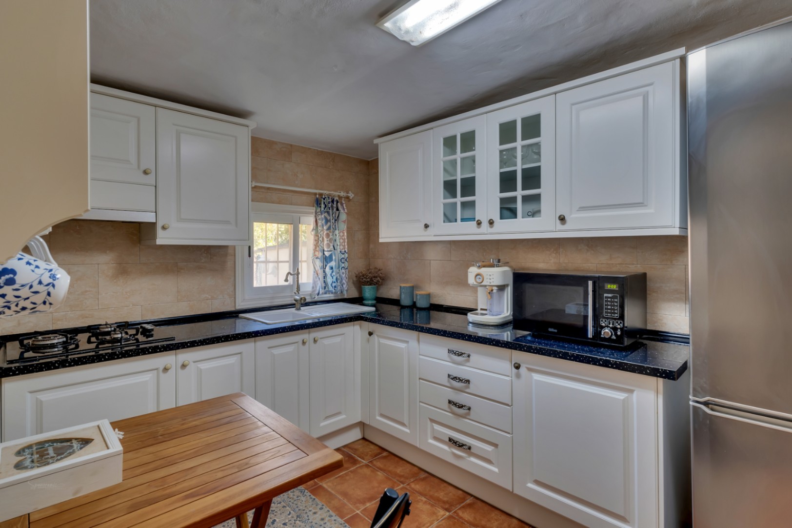 Cocina funcional con mobiliario blanco de estilo clásico, encimera oscura moteada, electrodomésticos de acero inoxidable y suelo de gres en tonos tierra.