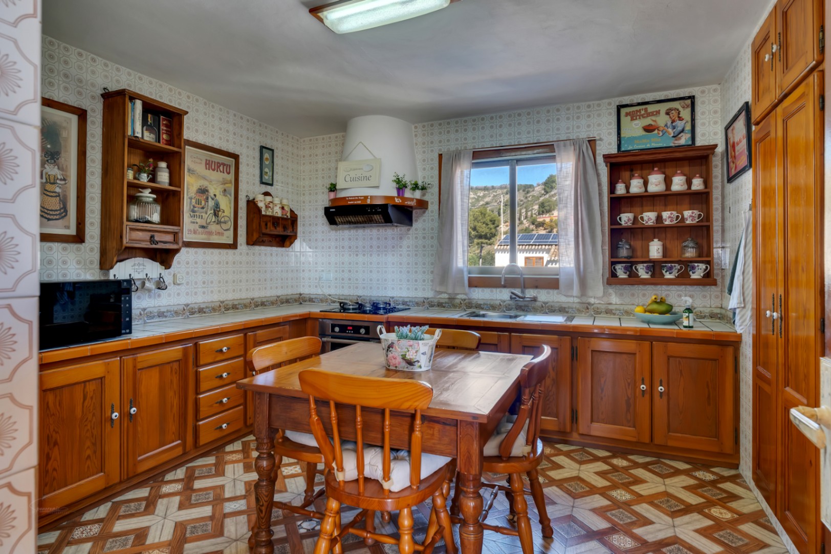 Cocina rústica con mobiliario de madera, mesa de comedor central y paredes alicatadas. Incluye una ventana con vistas despejadas a la montaña y suelos cerámicos decorativos.