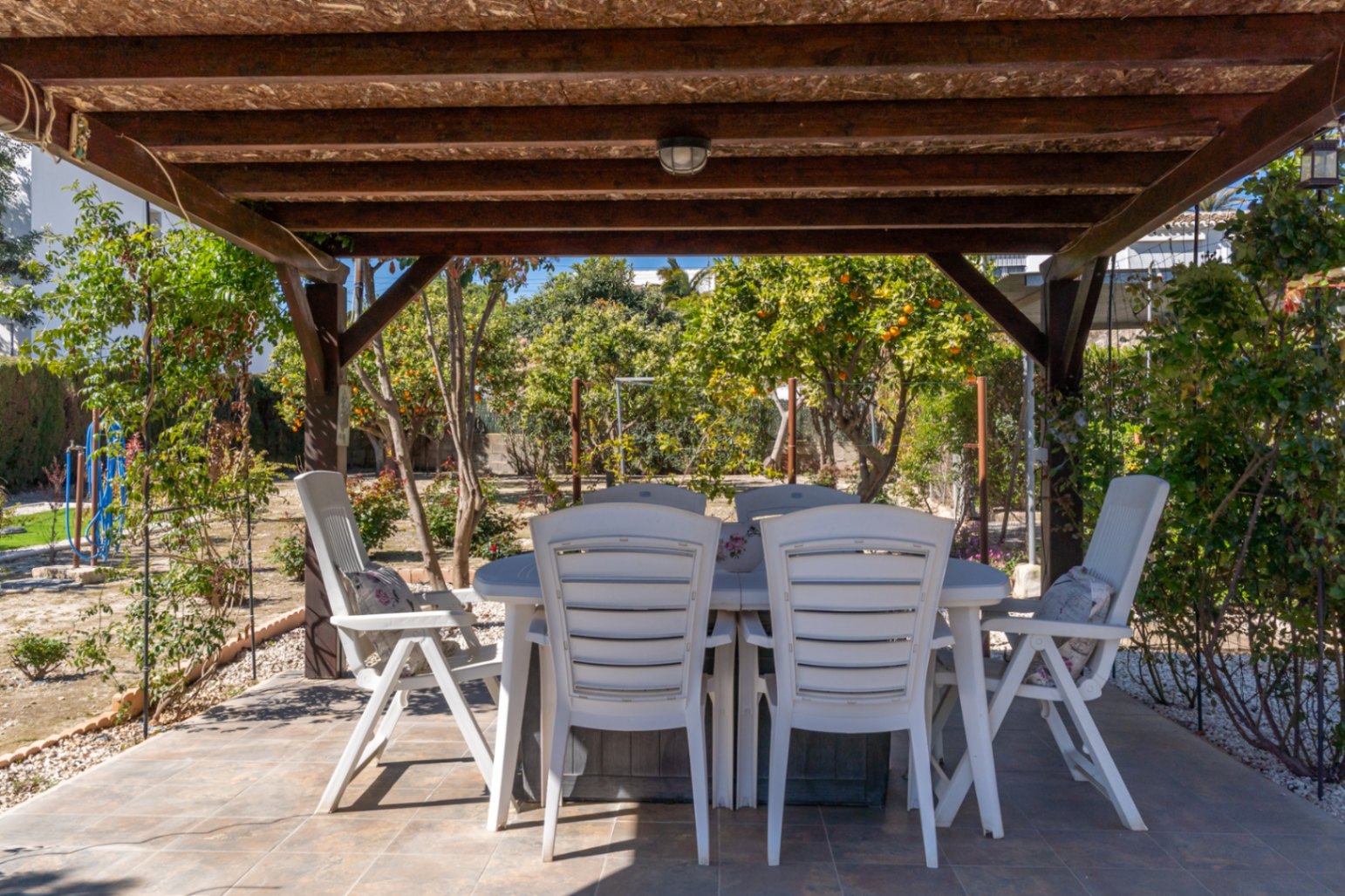 Comedor exterior bajo una pérgola de madera, rodeado de un jardín mediterráneo con árboles frutales y suelos de gres.