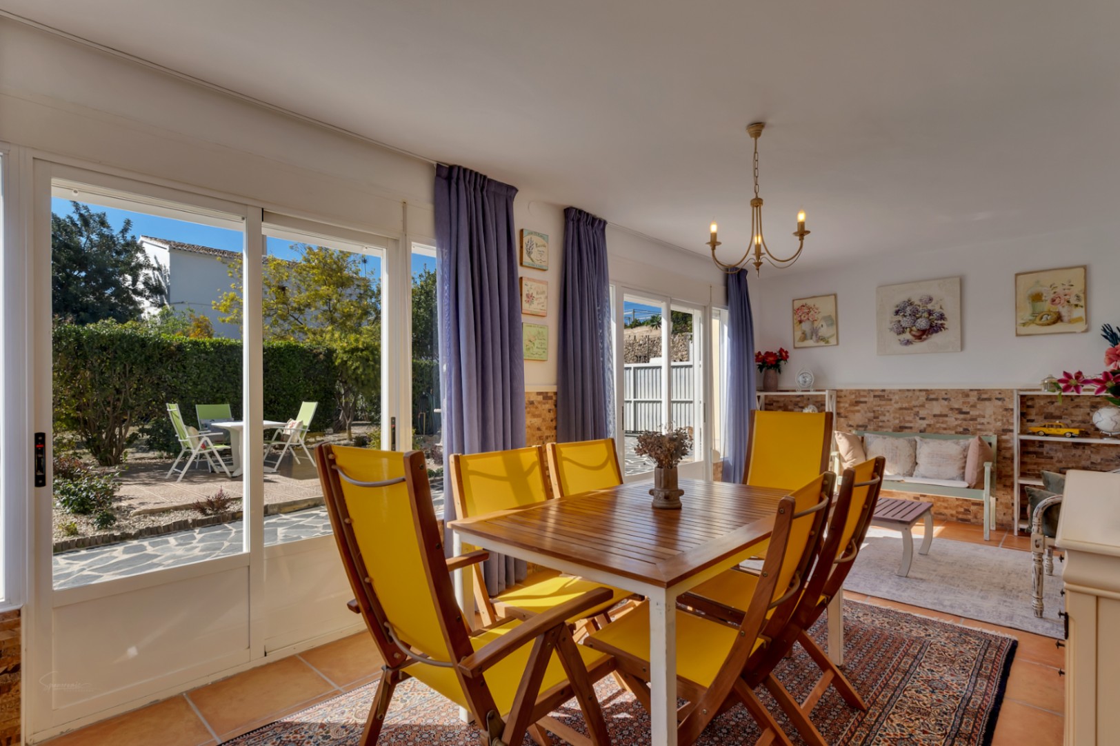 Comedor luminoso con mesa de madera y sillas amarillas, suelos de terracota y acceso directo al jardín a través de amplios ventanales correderos.