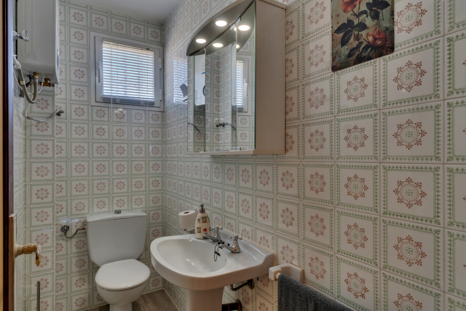 Cuarto de baño funcional con paredes alicatadas con motivos geométricos, lavabo de pedestal, inodoro blanco y armario de espejo con iluminación integrada.