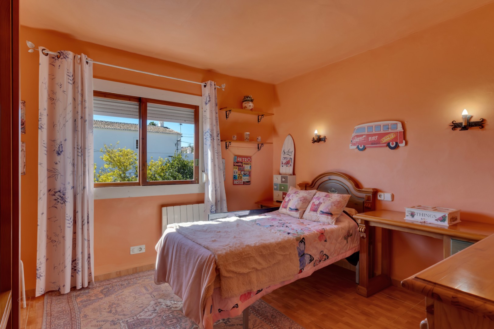 Dormitorio luminoso con paredes en tono melocotón, mobiliario de madera, escritorio integrado y ventana con vistas. Incluye calefacción por radiador y suelo cerámico.