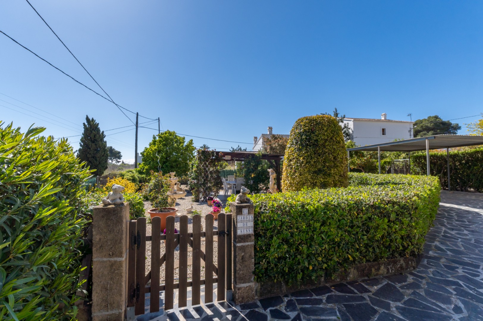 Entrada al jardín con puerta de madera, pilares de piedra y setos cuidados. Incluye zona de grava, pérgola al fondo y acceso al carport sobre suelo de piedra.