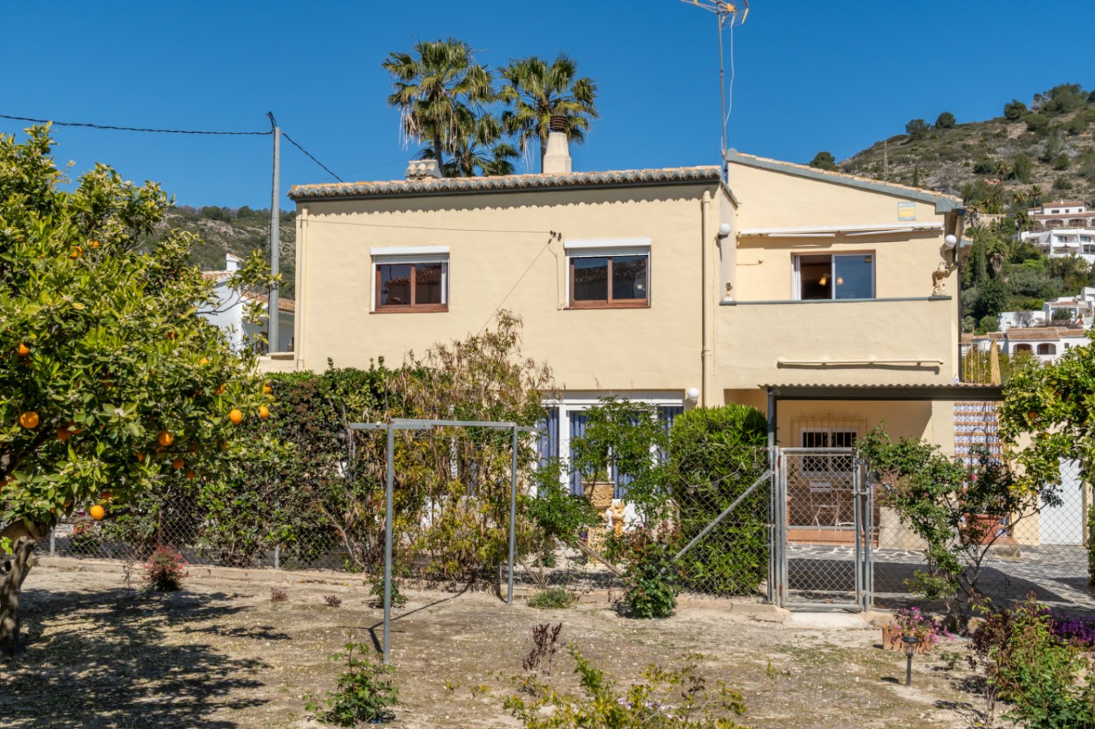 Fachada exterior de la villa con muros en tono crema, rodeada de un jardín con naranjos y palmeras bajo el cielo despejado de Jávea.