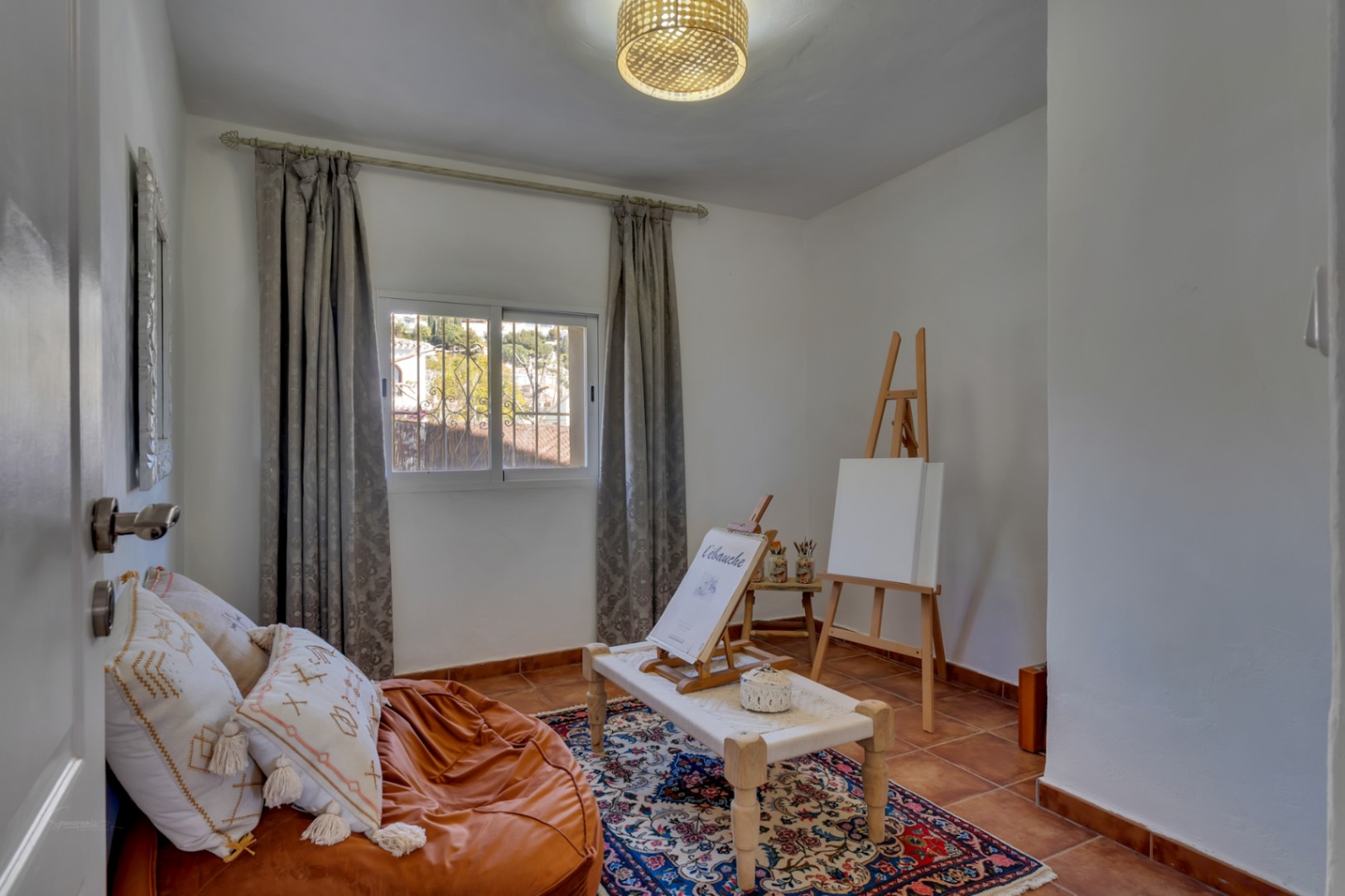 Habitación versátil con suelos de terracota, configurada como estudio de arte con caballete de madera, luz natural y textiles de estilo mediterráneo.