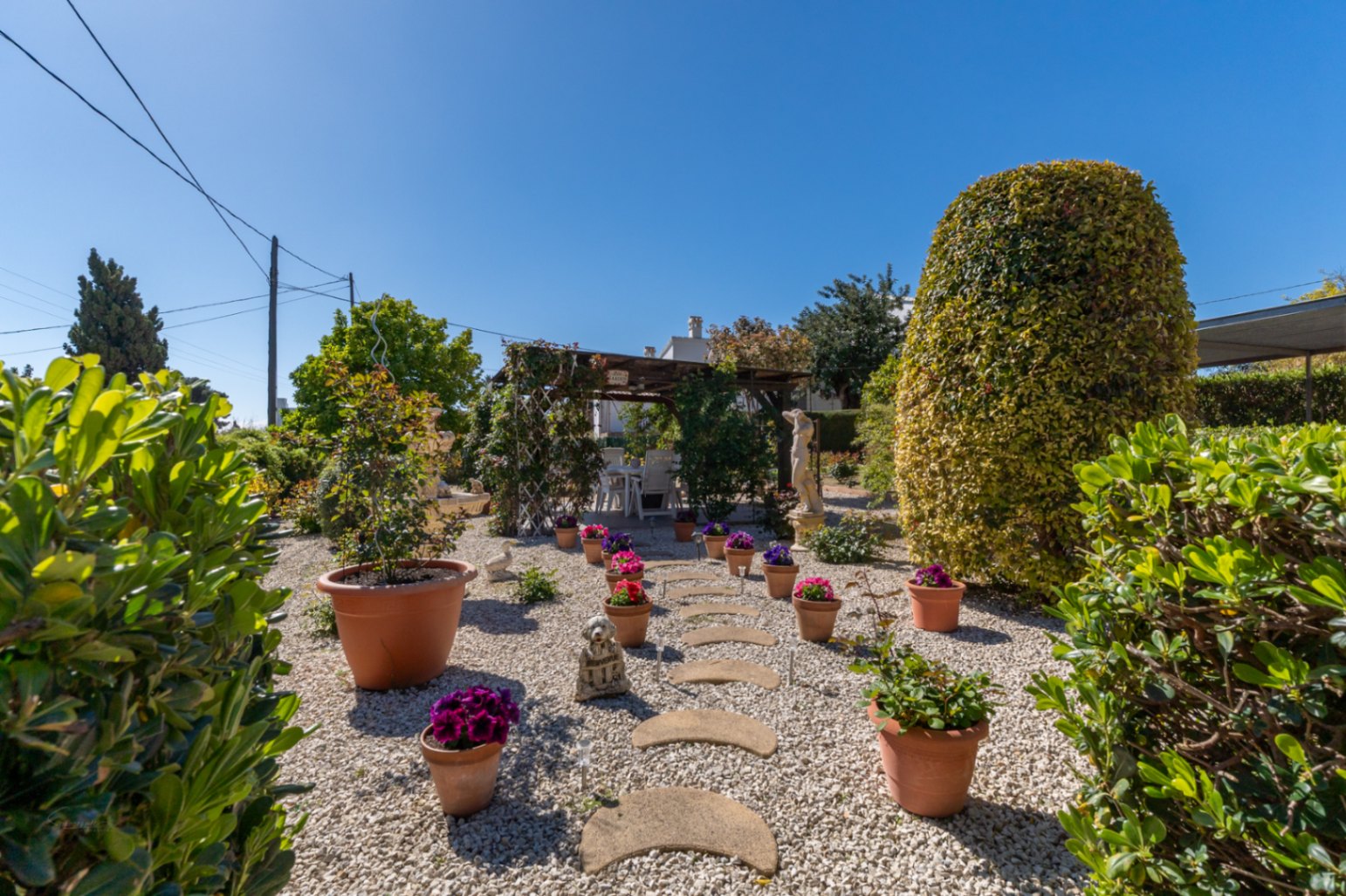 Jardín mediterráneo con suelo de grava, camino de piedras curvas y macetas de terracota. Al fondo, una pérgola de madera con zona de comedor exterior.