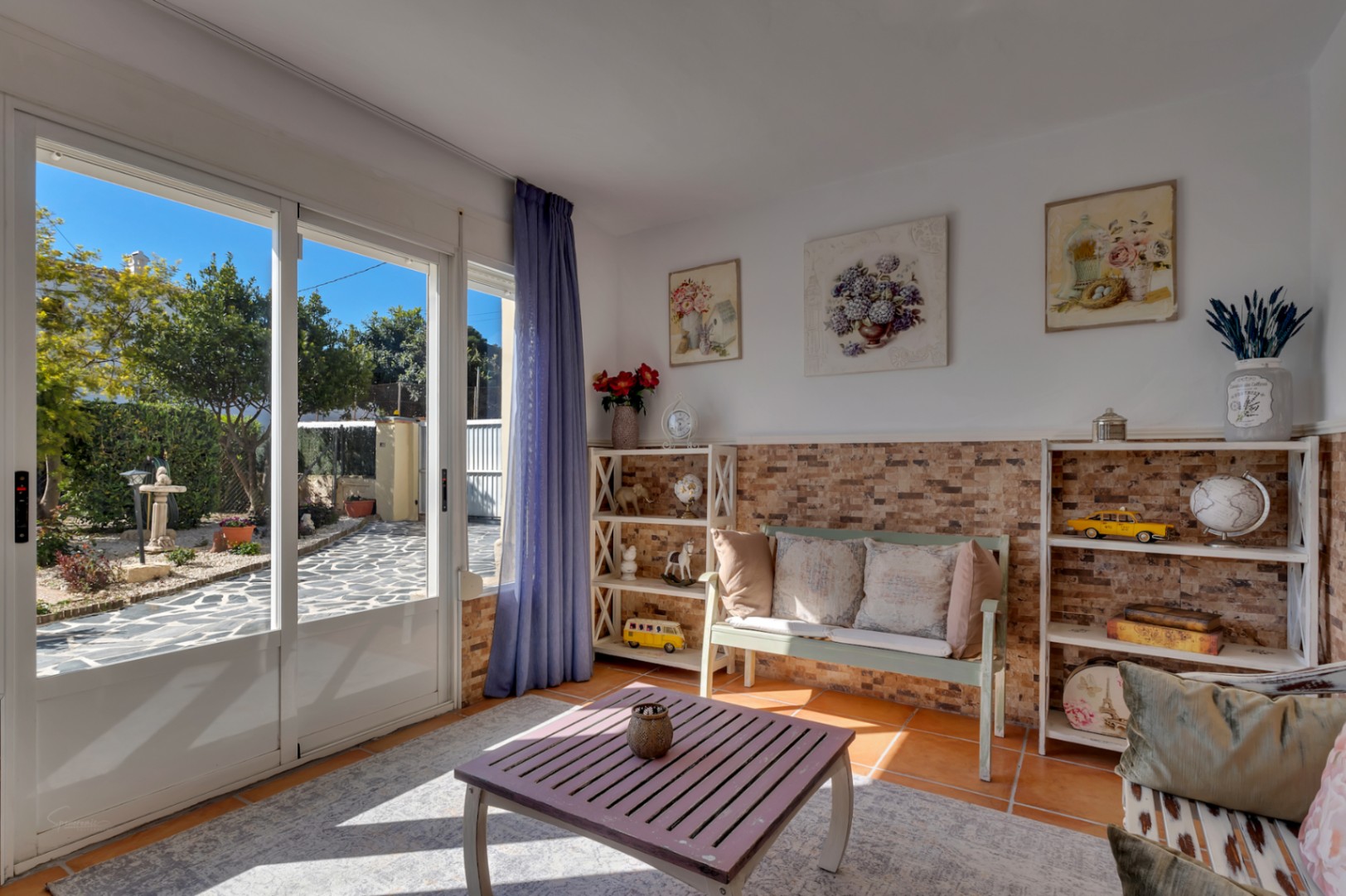 Sala de estar luminosa con acceso directo al jardín, paredes con zócalo de piedra decorativa, suelos de terracota y mobiliario de estilo rústico mediterráneo.