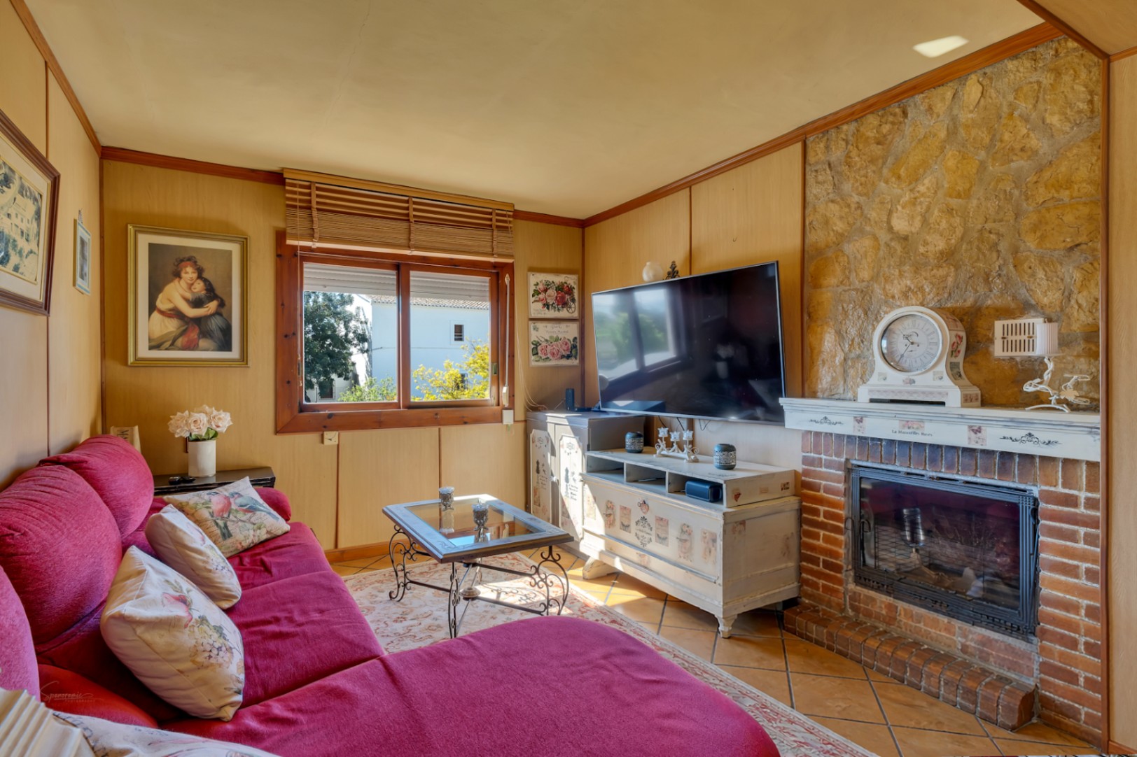 Salón acogedor con chimenea de ladrillo y piedra natural, paredes revestidas en madera y un amplio sofá rojo que define la zona de estar.