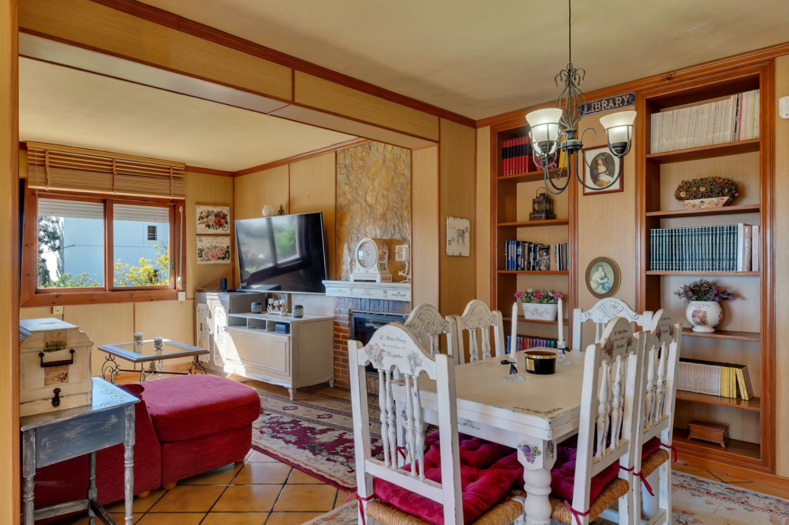 Salón comedor con chimenea de piedra, paredes paneladas en madera y estanterías integradas. Incluye mobiliario de estilo rústico y suelos de gres cerámico.