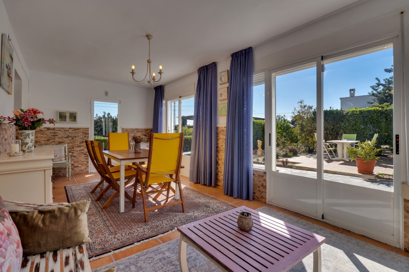 Salón comedor luminoso con acceso directo al jardín, mesa de comedor con sillas amarillas y amplias puertas correderas de cristal que integran el exterior.