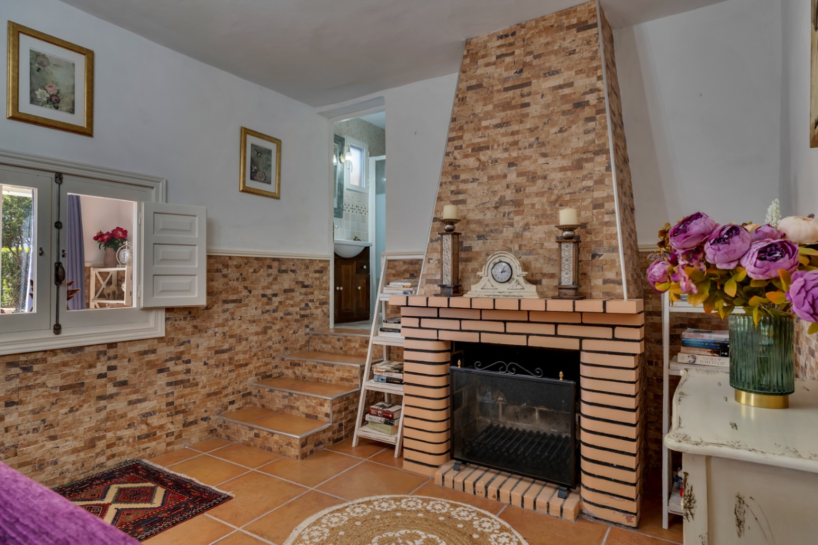 Salón con chimenea de obra revestida en piedra, suelos de terracota y acceso mediante escalones a un baño. Incluye detalles rústicos y una ventana interior con contraventanas blancas.