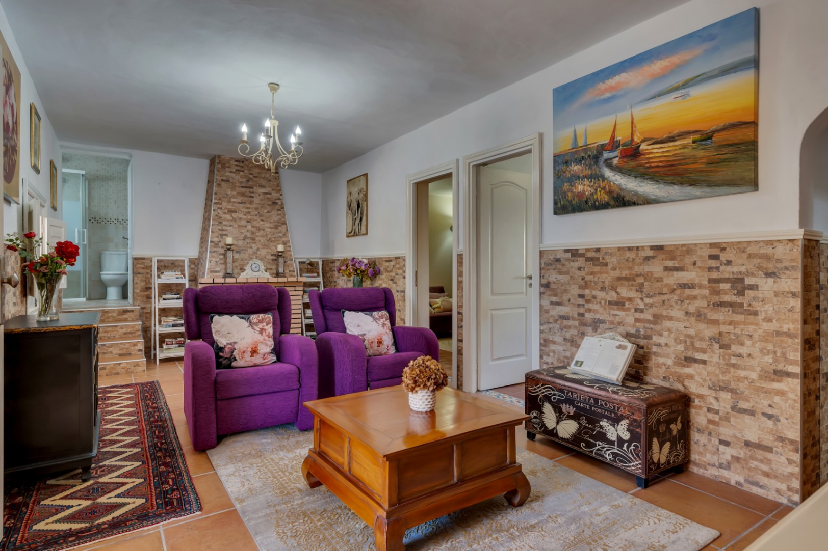 Salón con chimenea revestida en piedra, sillones violetas y suelos cerámicos. Incluye acceso a estancias adicionales y un baño completo visible al fondo.