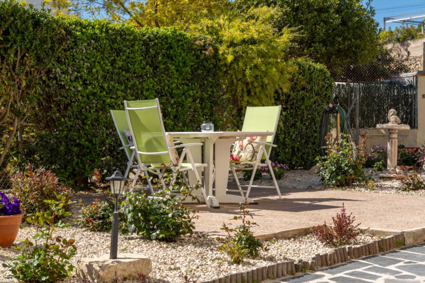 Zona de estar exterior pavimentada con mesa blanca y sillas verdes, rodeada de grava decorativa, setos altos de privacidad y vegetación mediterránea cuidada.