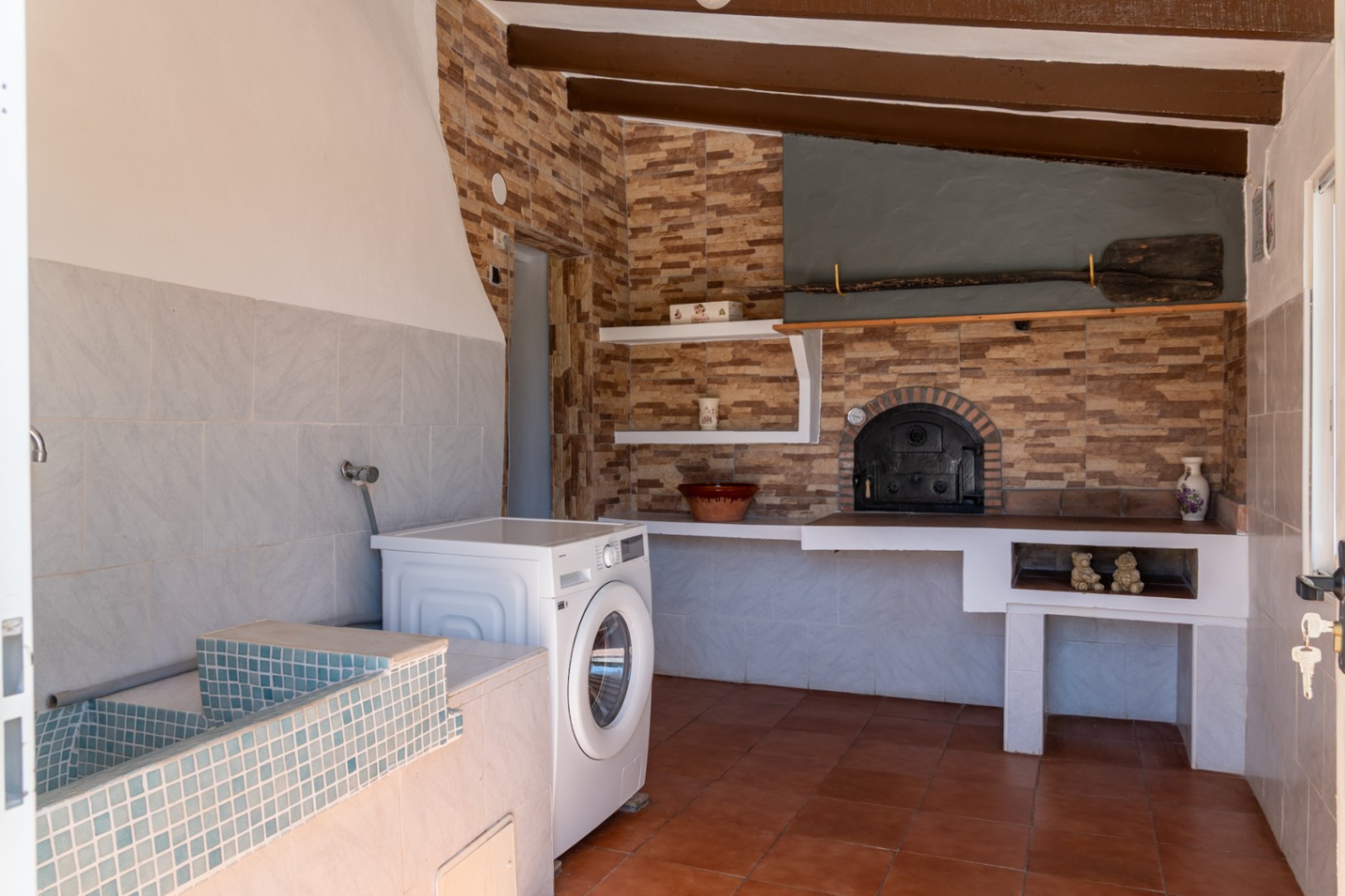 Zona de lavandería y cocina rústica con horno de leña tradicional, fregadero de mosaico azul, lavadora y techos con vigas de madera vistas.