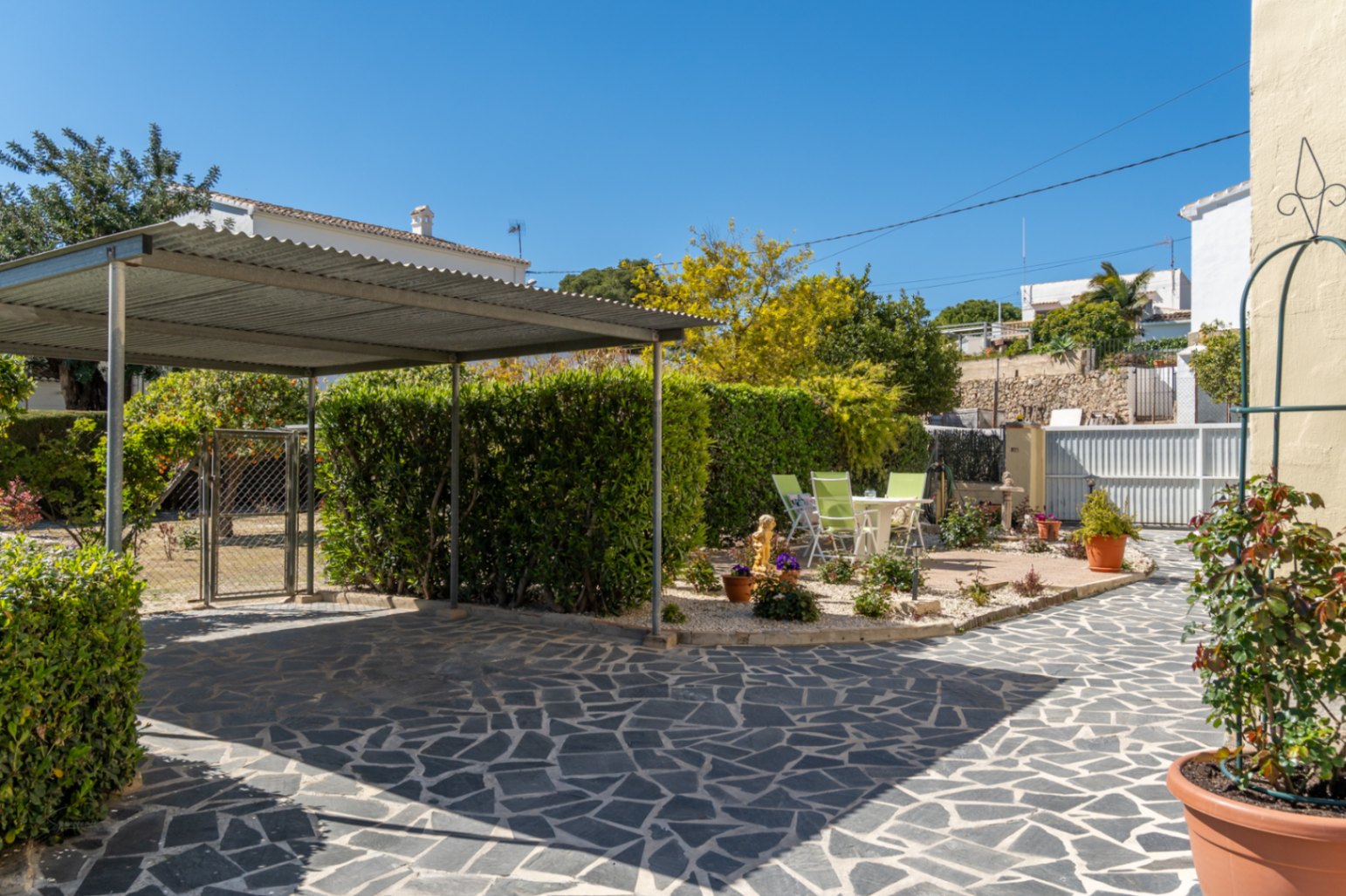 Zona exterior con pavimento de piedra irregular, carport metálico cubierto y un área de descanso rodeada de setos cuidados y vegetación mediterránea.