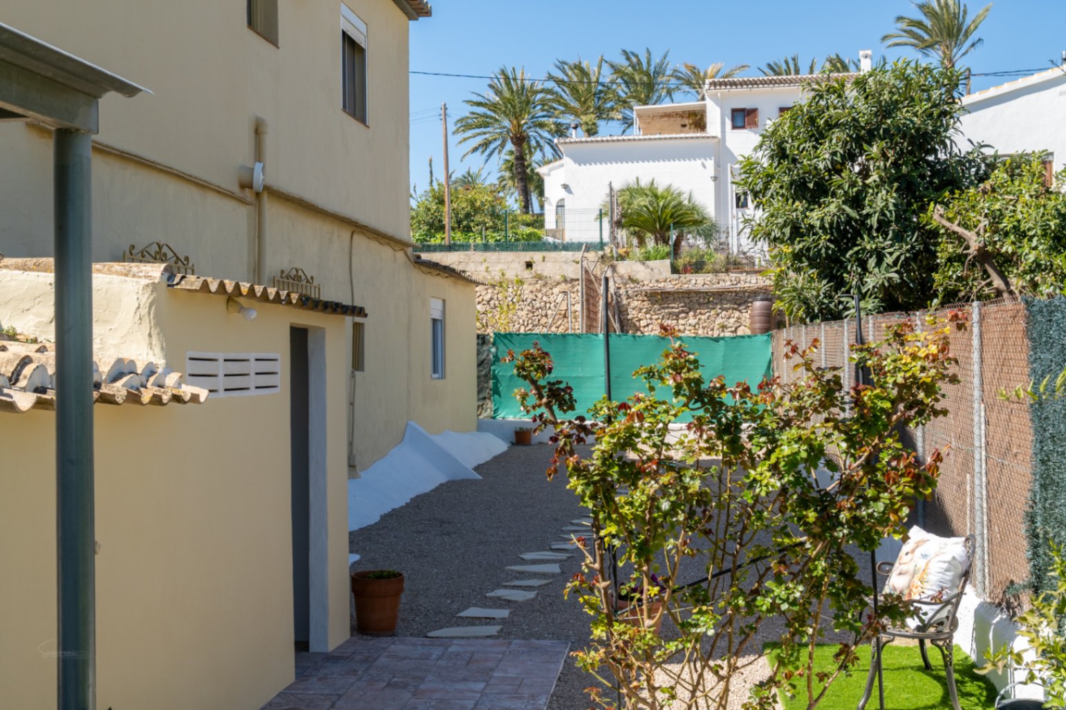 Zona exterior lateral con camino de grava y piedras, pequeña área de césped con mobiliario y muros de piedra tradicionales bajo el sol mediterráneo.