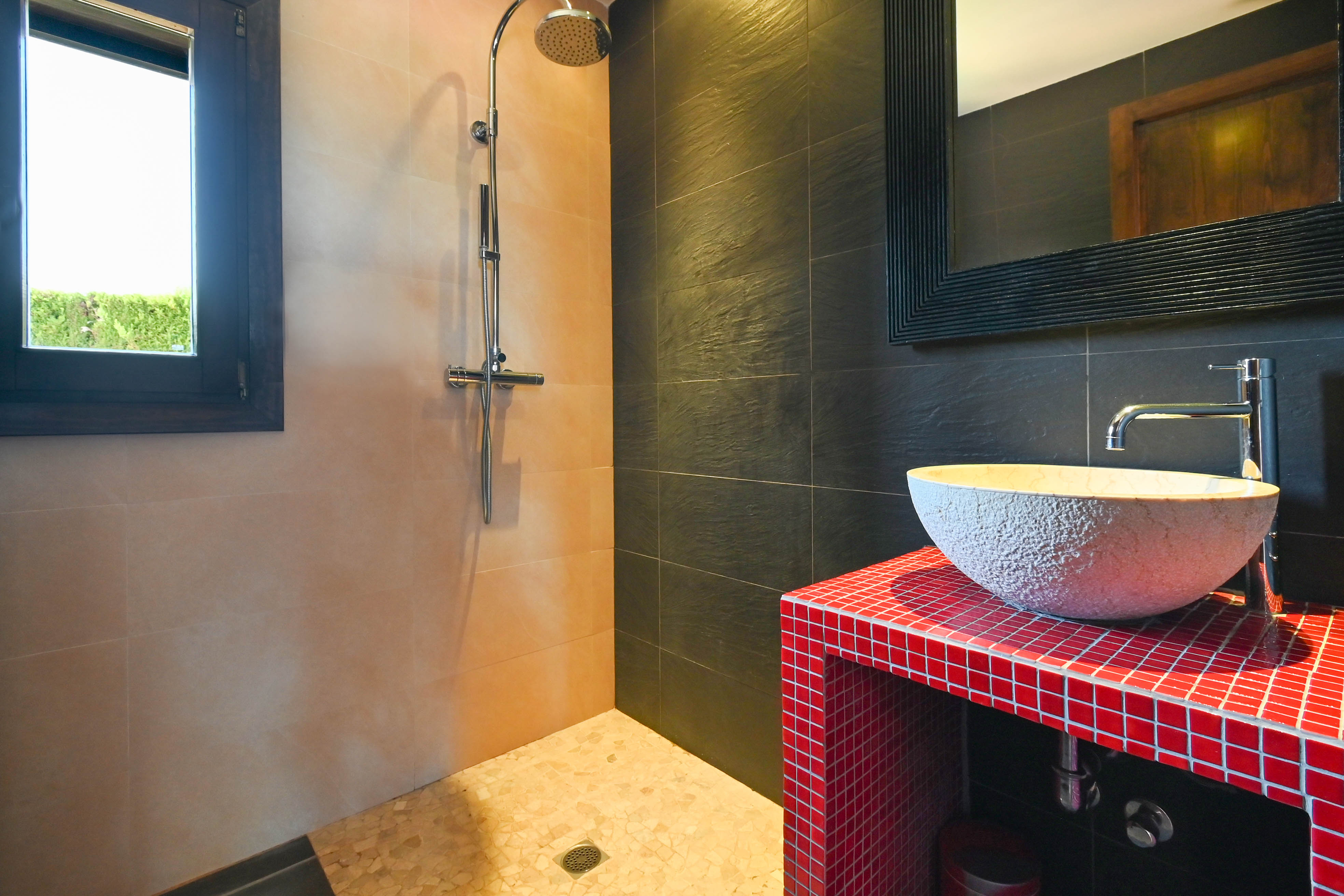 Detalle de baño con lavabo rústico sobre encimera de mosaico rojo y ducha con azulejos color arena.
