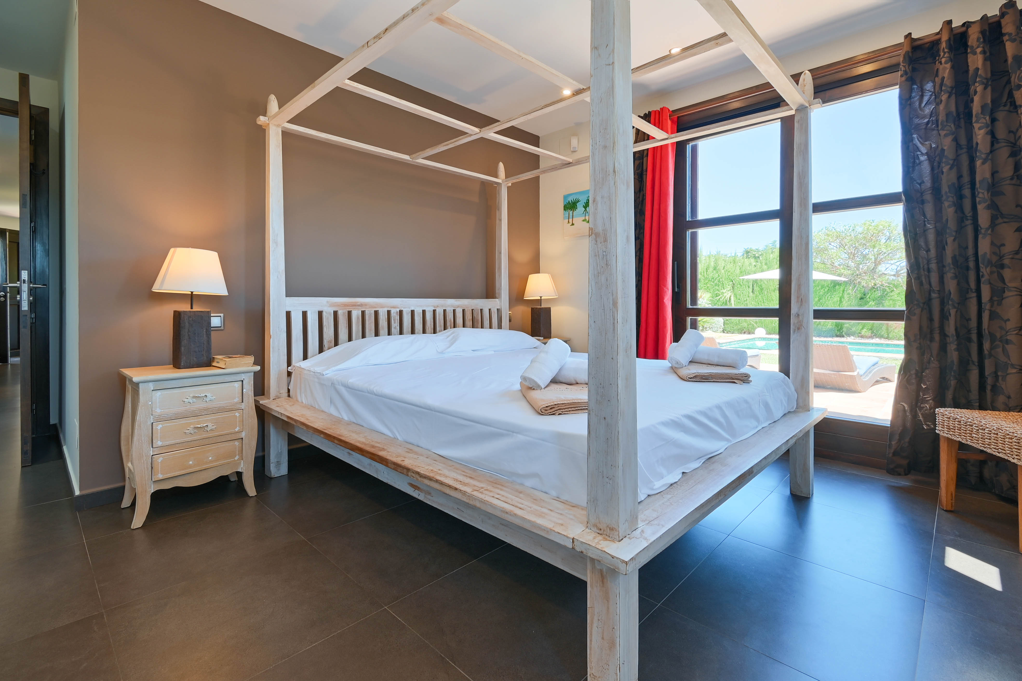 Dormitorio con cama con dosel de madera blanca, mesita de noche decapada y vista a la piscina.