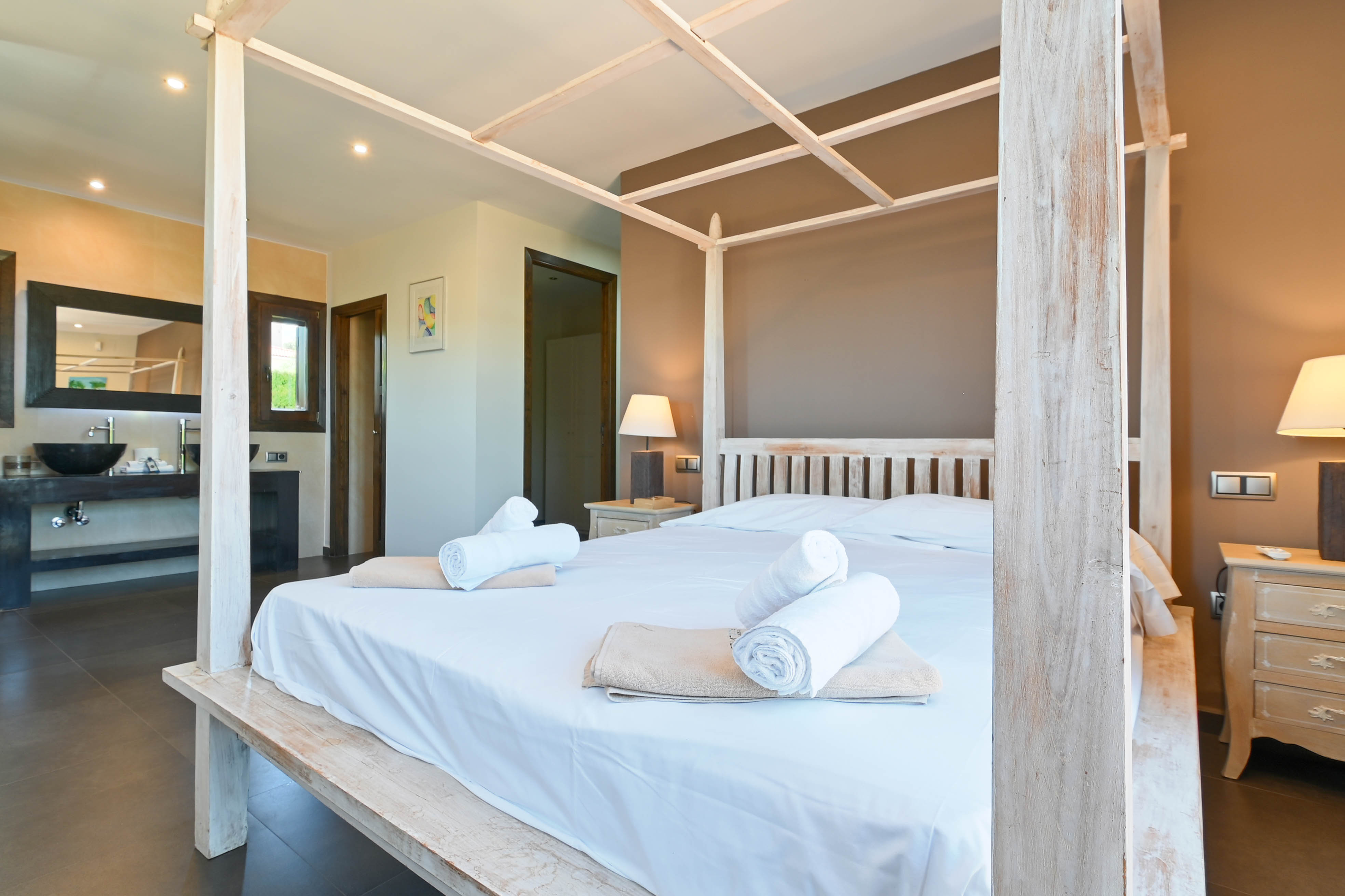 Dormitorio con cama con dosel de madera blanca, toallas enrolladas y baño con lavabo moderno.