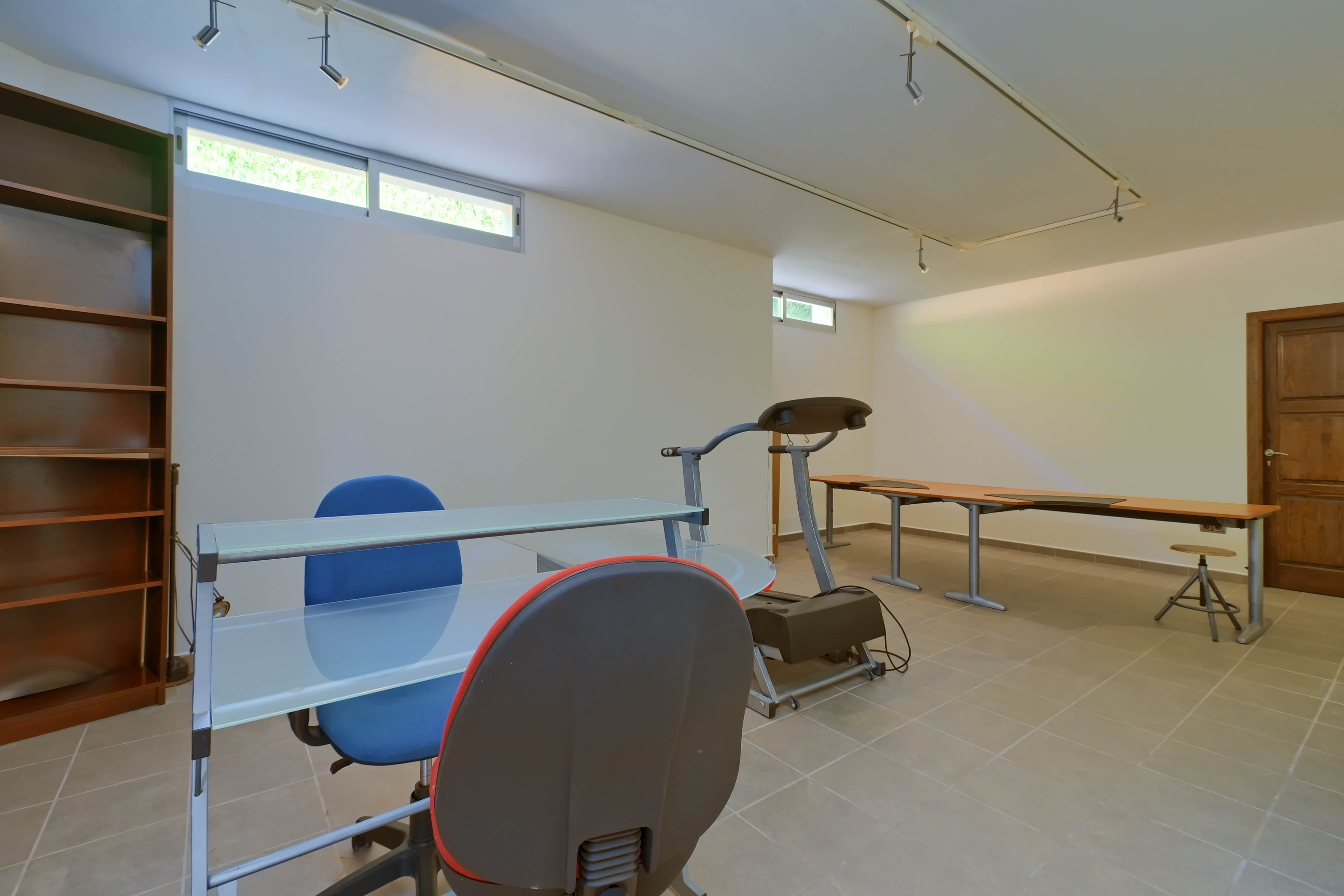 Espacio funcional con estantería, escritorio de cristal y cinta de correr en baldosa neutra.