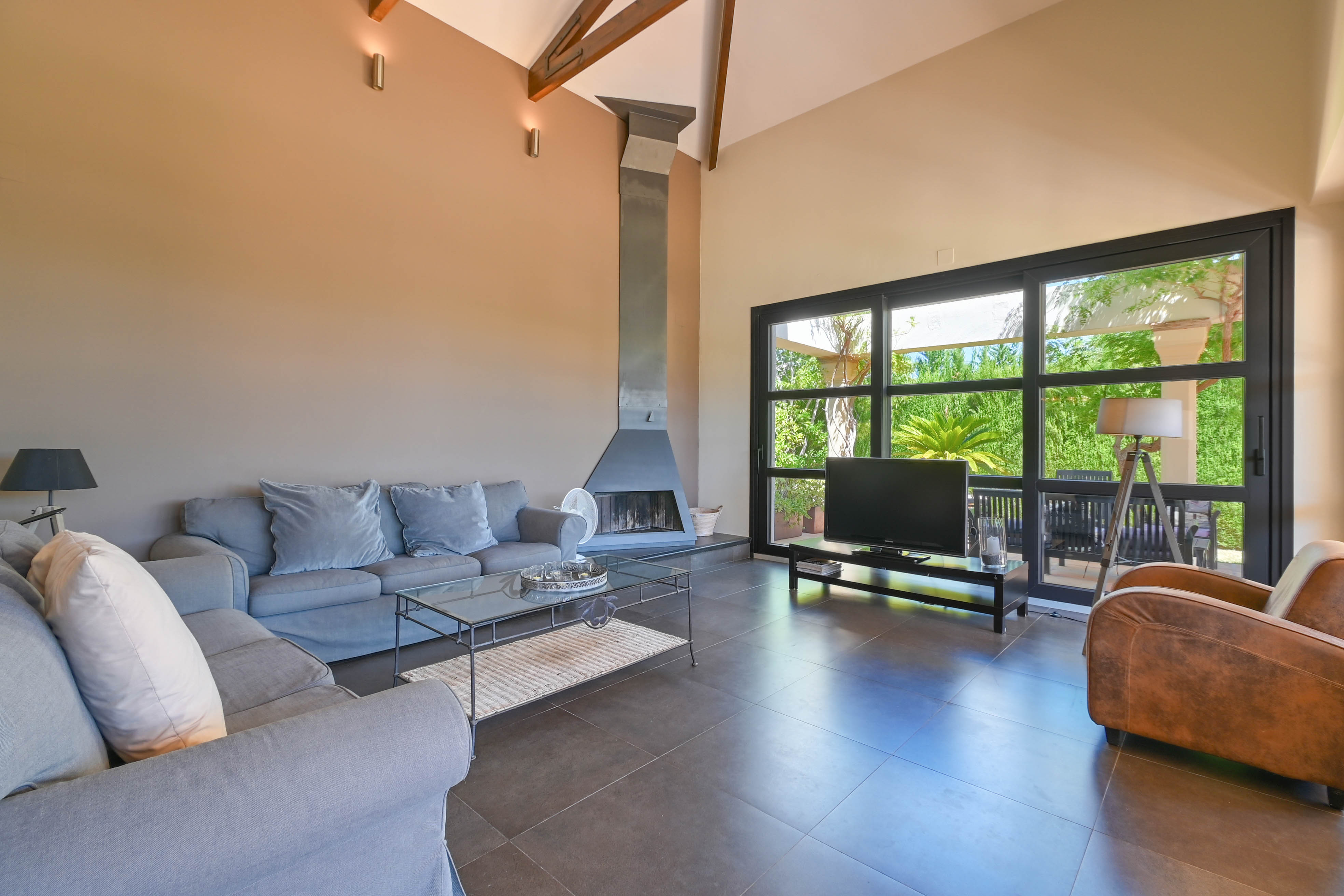 Salón amplio con chimenea metálica, sofá gris y ventanales al jardín en Denia, Costa Blanca.