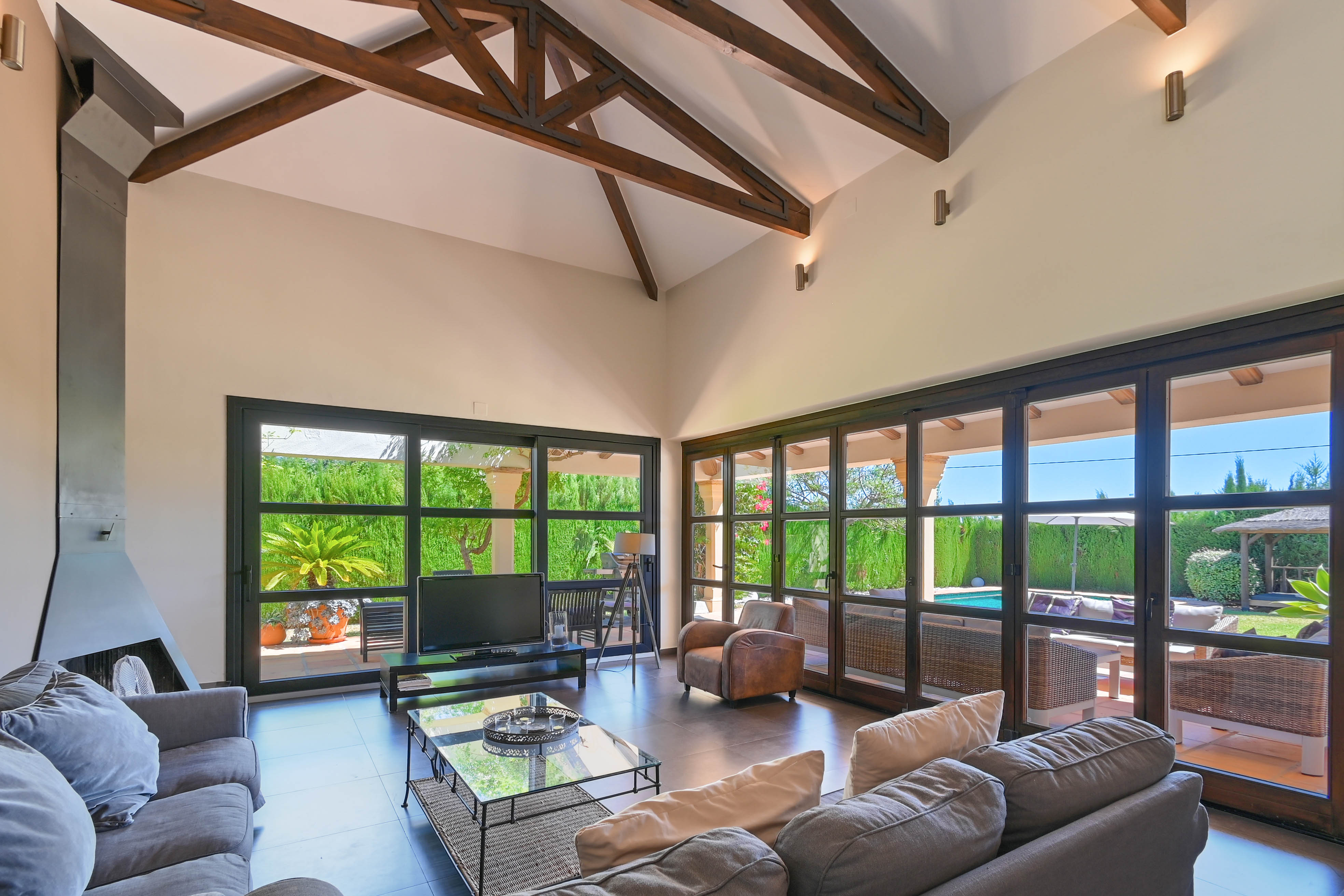 Salón luminoso con vigas de madera, chimenea moderna y vistas al jardín con piscina a través de amplios ventanales.
