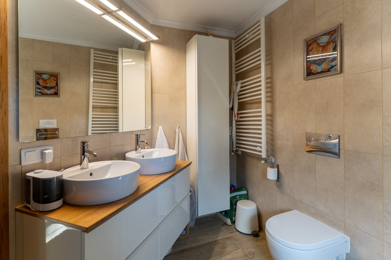 Baño con doble lavabo sobre encimera de madera, toallero calefactado y alicatado cerámico color arena.