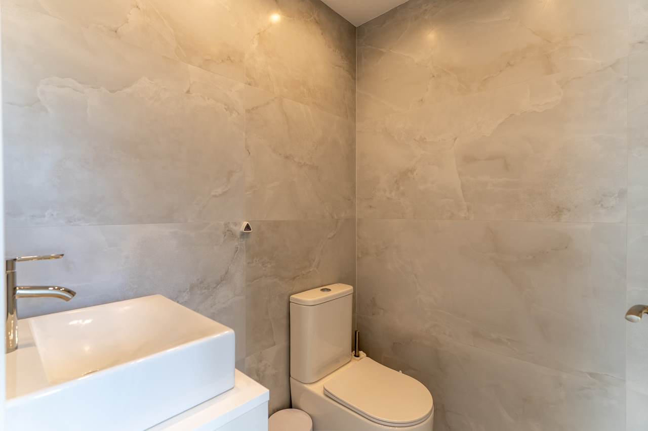 Detalle de baño moderno con paredes de baldosa veteada, lavabo cuadrado sobre encimera blanca y sanitarios.