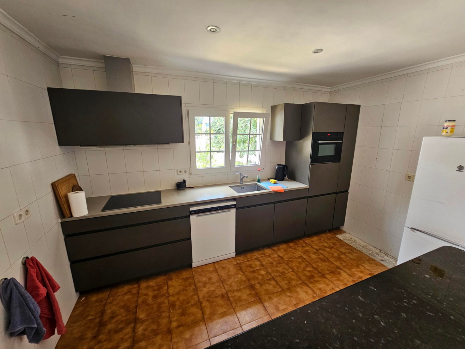 Cocina con muebles grises, encimera clara y baldosas blancas. Ventana con vistas al exterior.