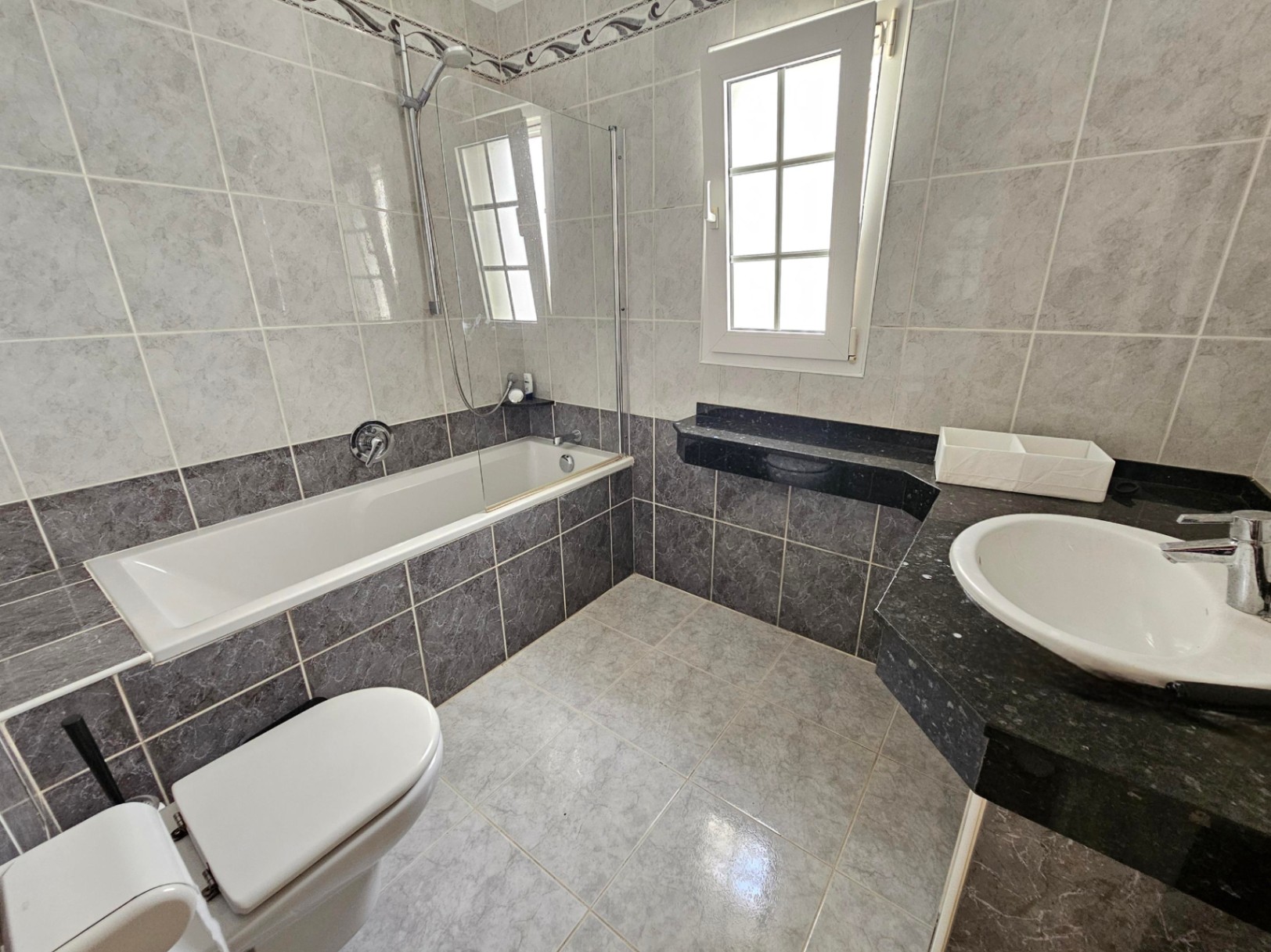 Cuarto de baño con azulejos grises, bañera con mampara y encimera de granito negro.