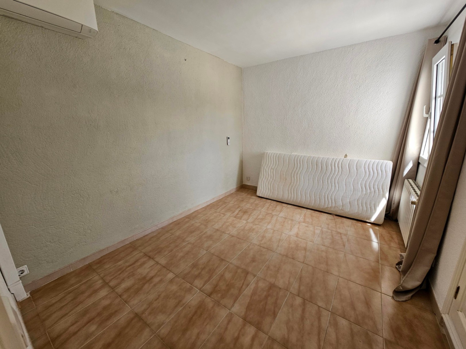 Habitación vacía con suelo de baldosas color crema y paredes texturizadas. Radiador visible bajo la ventana.