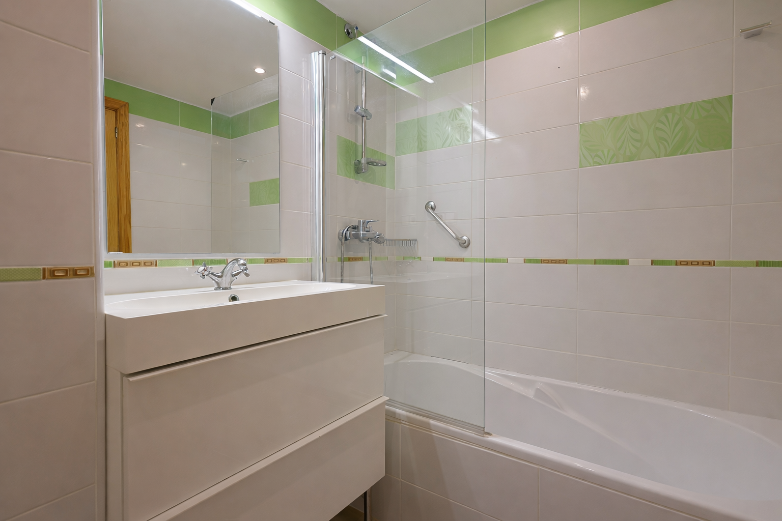 Baño con lavabo moderno, azulejos blancos y detalles verdes, y ducha con mampara de cristal.