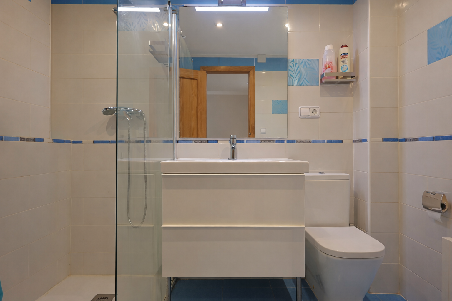 Cuarto de baño con azulejos blancos y detalles azules, espejo, lavabo moderno y ducha con mampara de cristal.