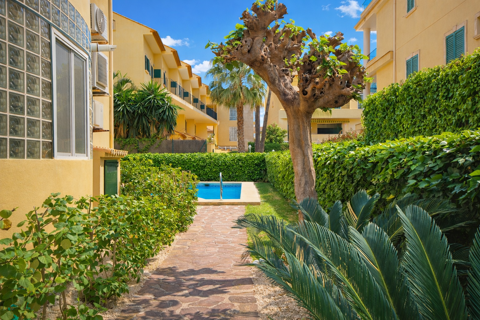 Sendero adoquinado que conduce a una piscina, rodeado de jardines y edificios amarillos.