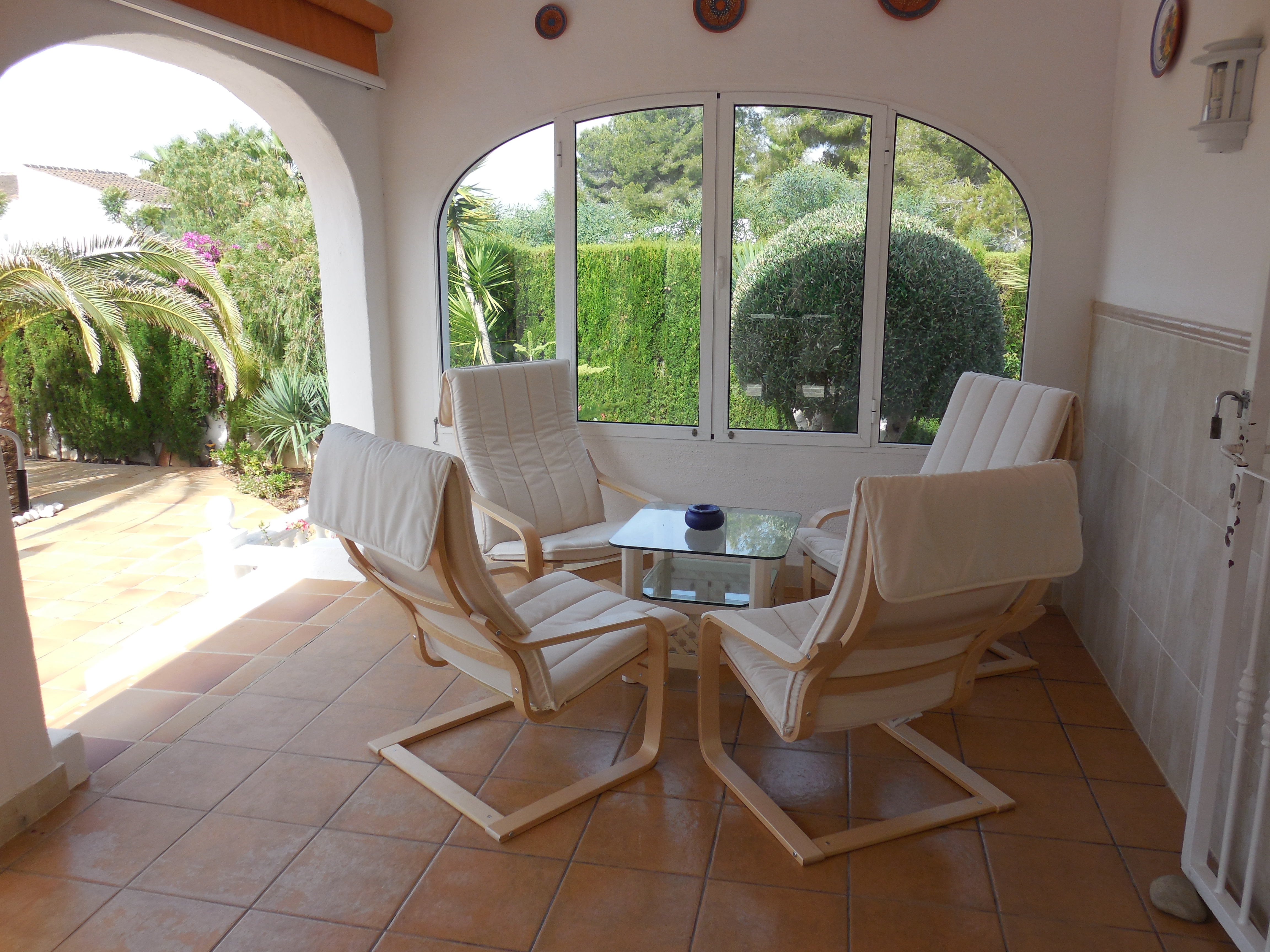 Terraza cubierta con cuatro sillones de madera clara y cojines blancos, mesa de cristal y vistas al jardín.