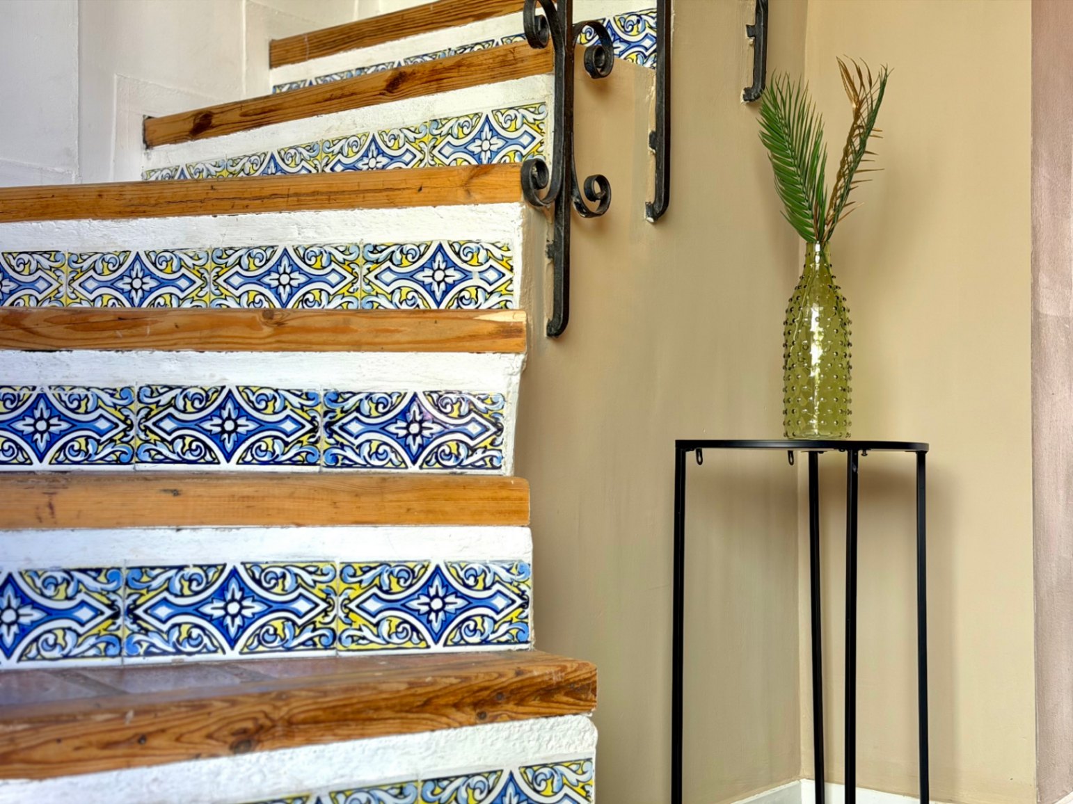 Detalle de escalera con azulejos decorativos y barandilla de hierro forjado. Jarrón verde con planta.
