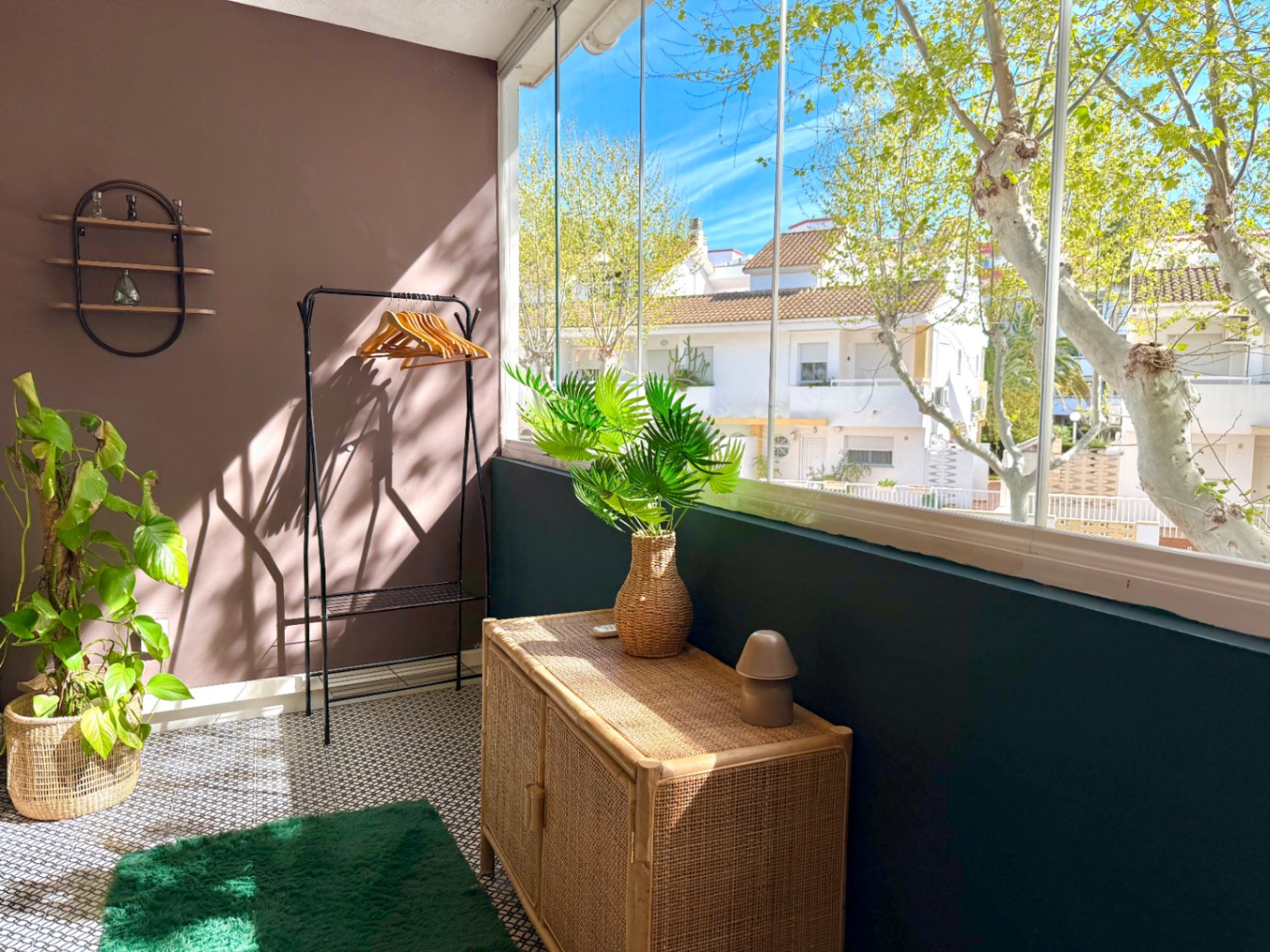 Detalle de interior con paredes en marrón y verde, mueble de mimbre y vistas a la calle desde la terraza acristalada.