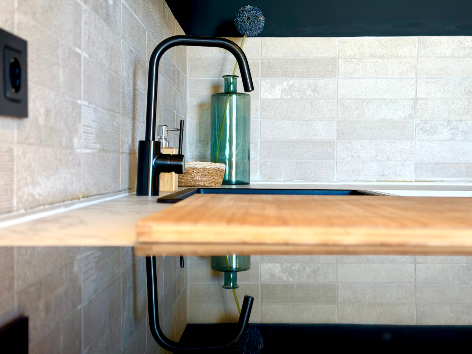 Detalle de la cocina con grifo negro, tabla de cortar de madera y revestimiento cerámico texturizado.