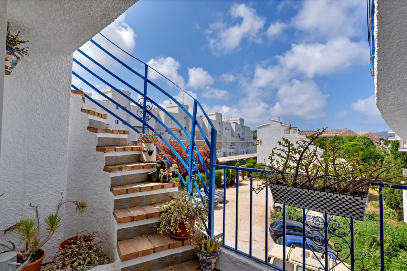 Escalera exterior con barandilla azul y detalles florales, que conduce a una terraza con vistas al aparcamiento.