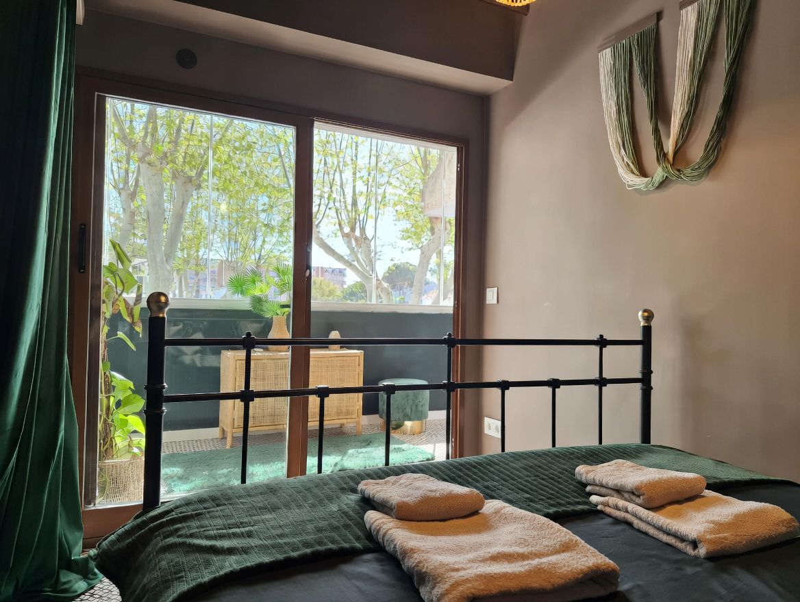 Habitación con cama de hierro forjado, balcón con vistas arboladas y decoración moderna en tonos neutros y verdes.