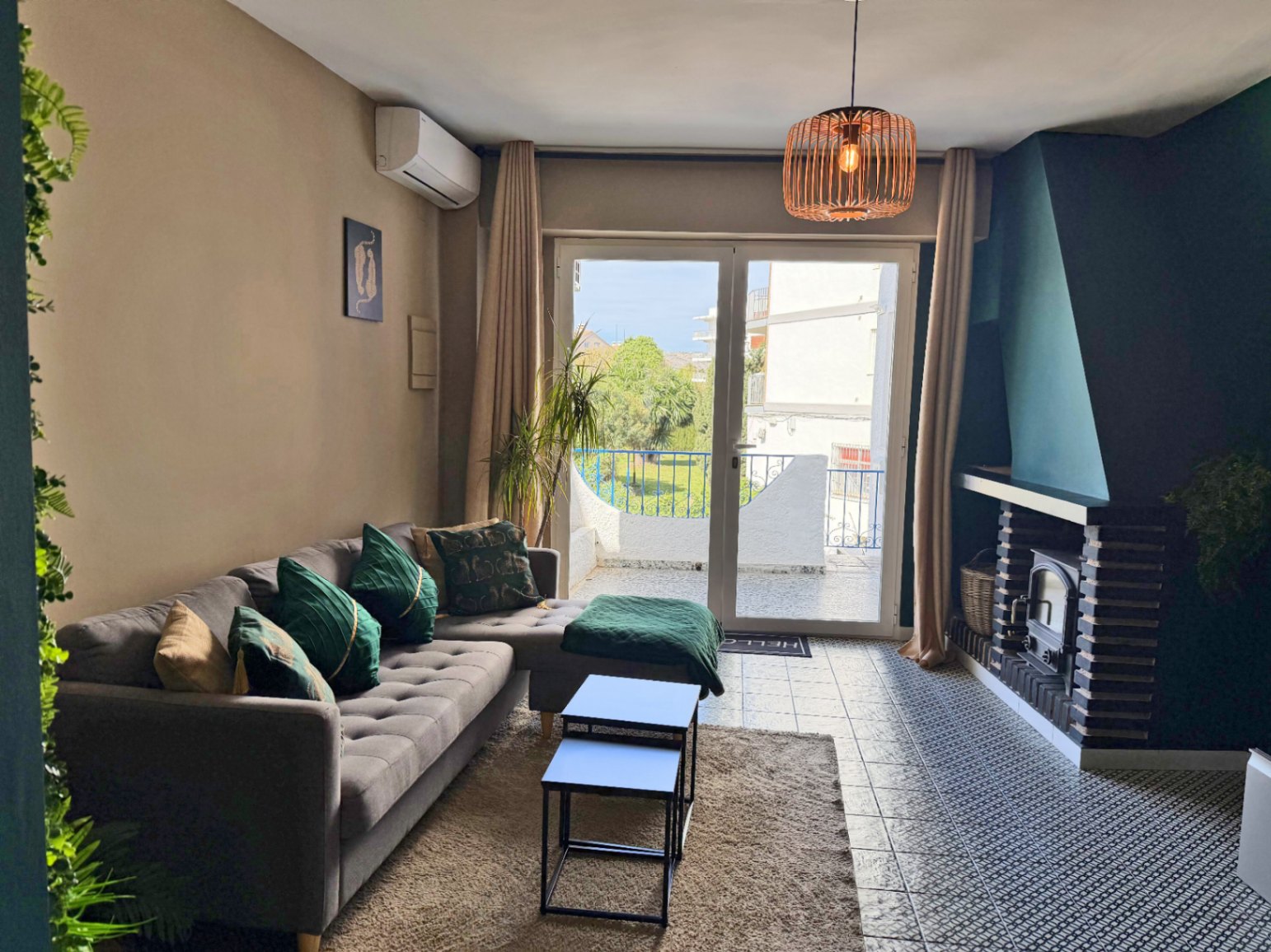 Salón con chimenea de ladrillo, sofá gris con cojines verdes y terraza con vistas.