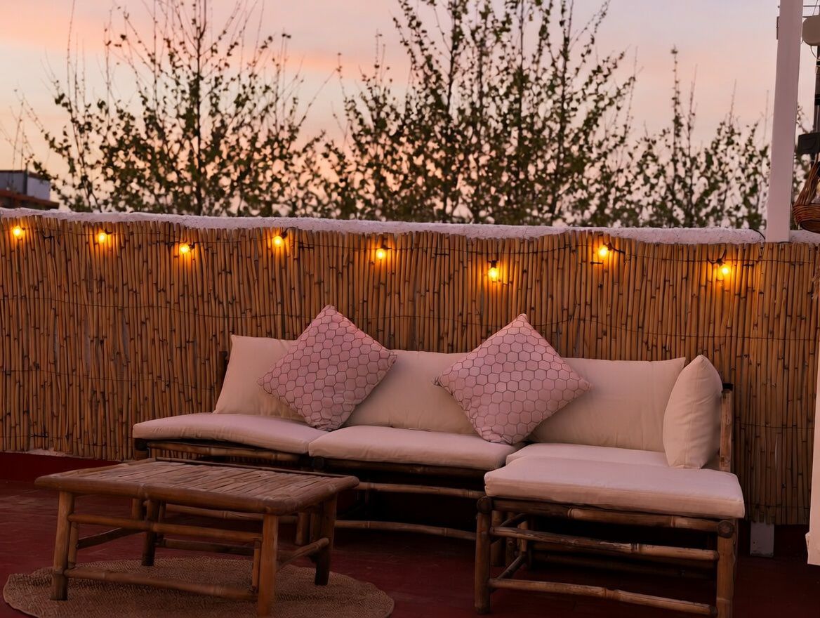 Terraza con sofá de bambú y cojines estampados, iluminada con luces cálidas, con vista al cielo al atardecer.
