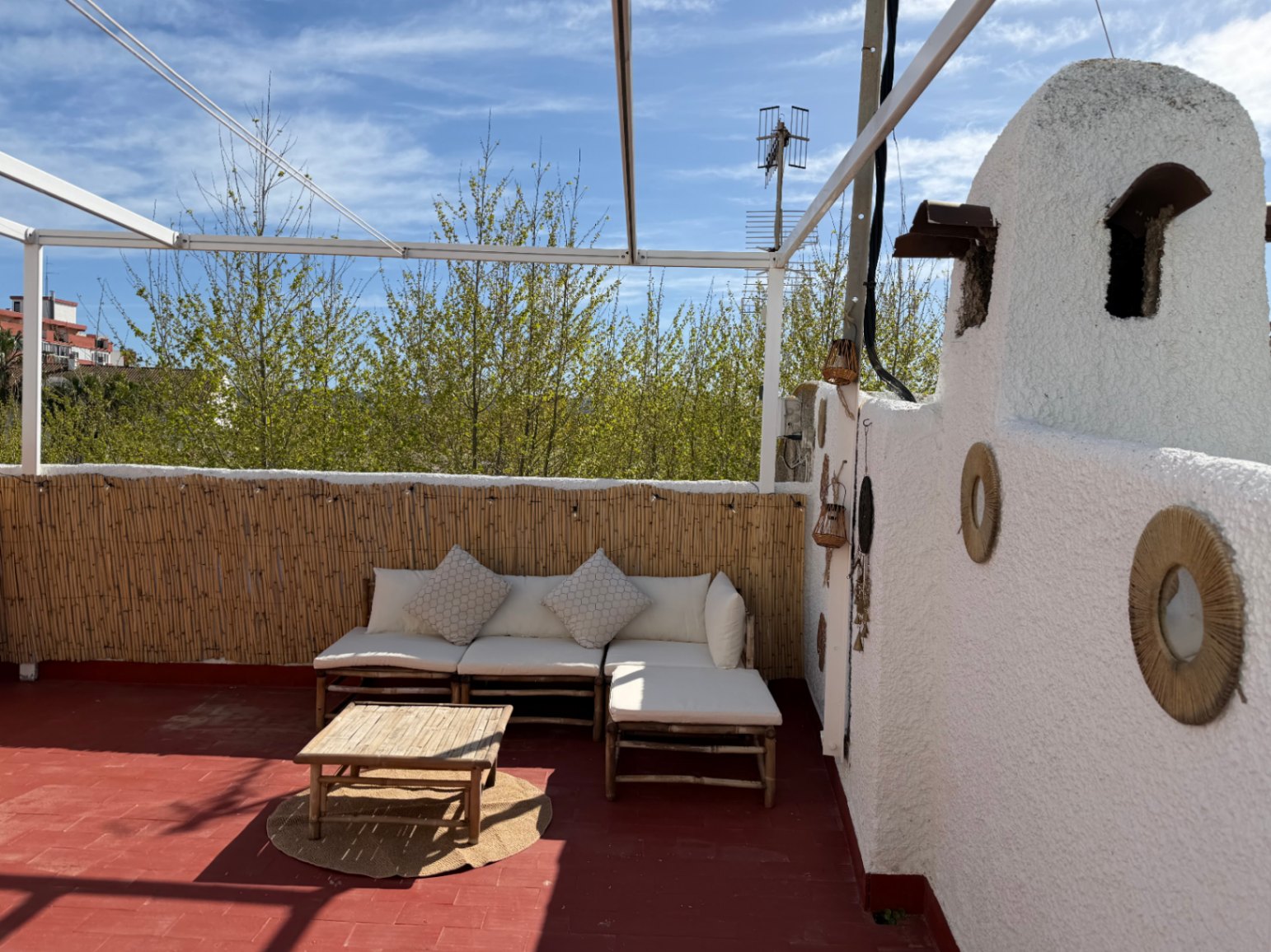 Terraza con suelo rojo, pared blanca decorada, sofá de bambú y vistas a la vegetación.